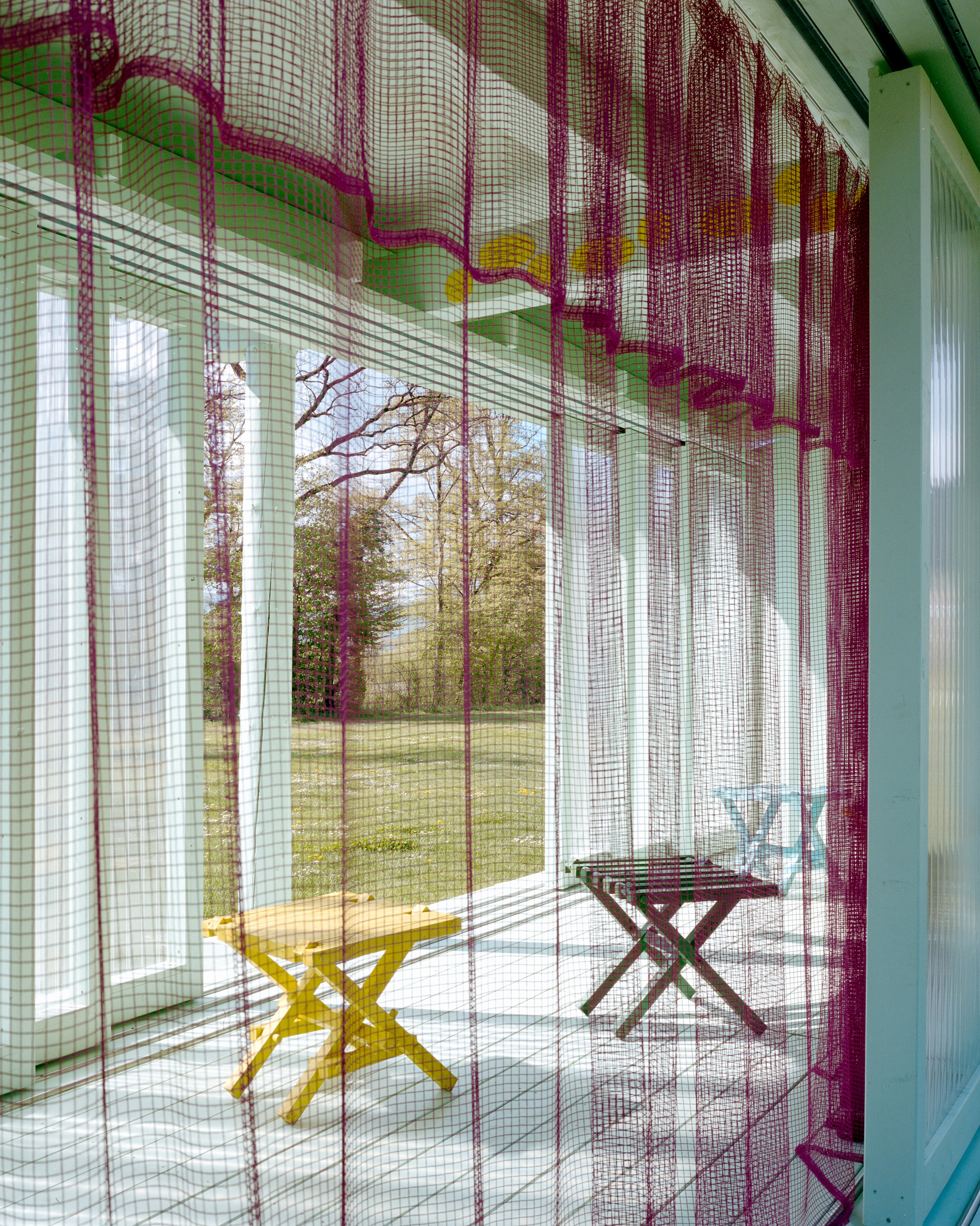 Magenta mesh curtain in the foreground with yellow and maroon folding stools on the porch floor, spring lawn and bare trees beyond