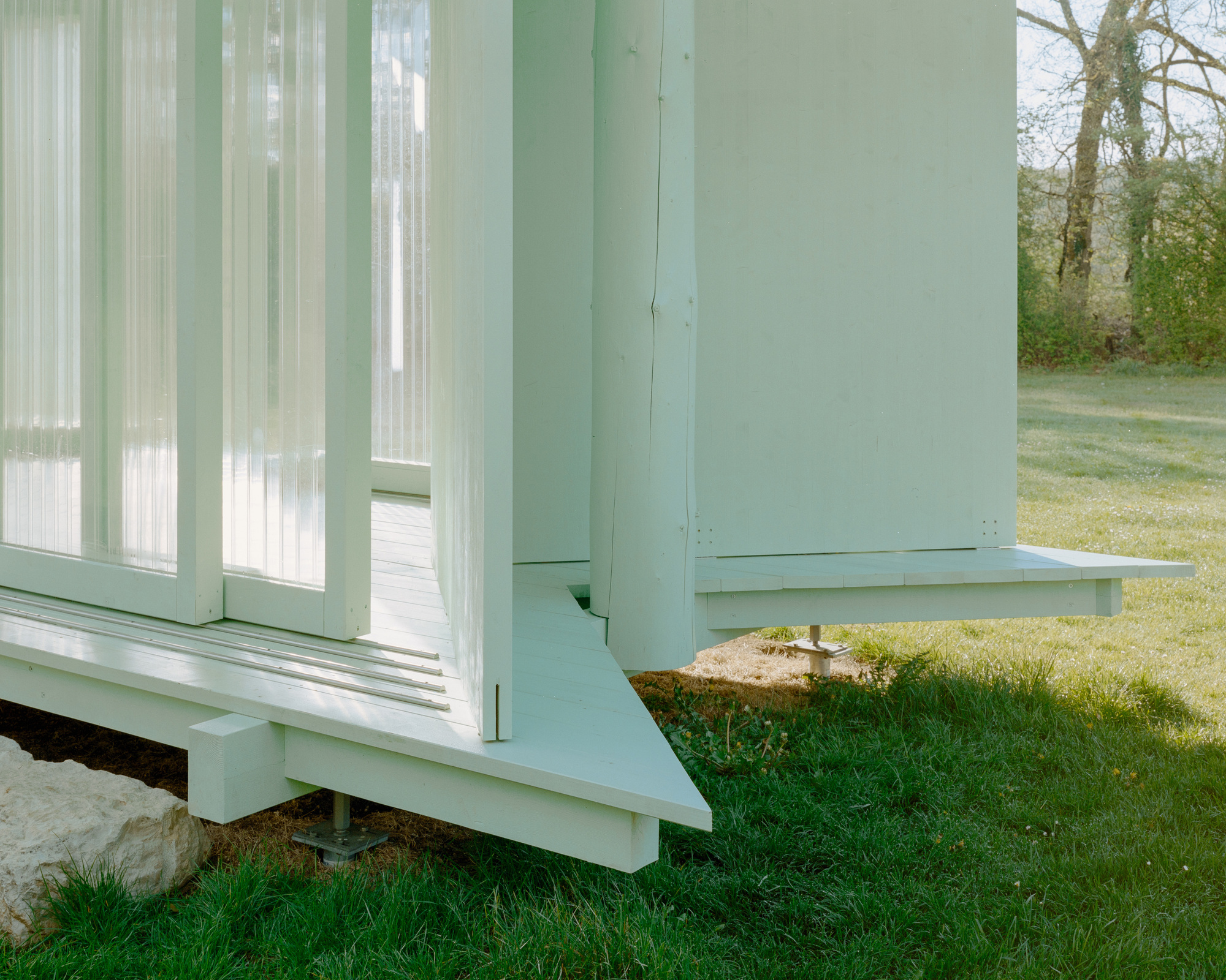 Corner of the raised timber deck on adjustable steel feet, with translucent panels and a log column over spring grass