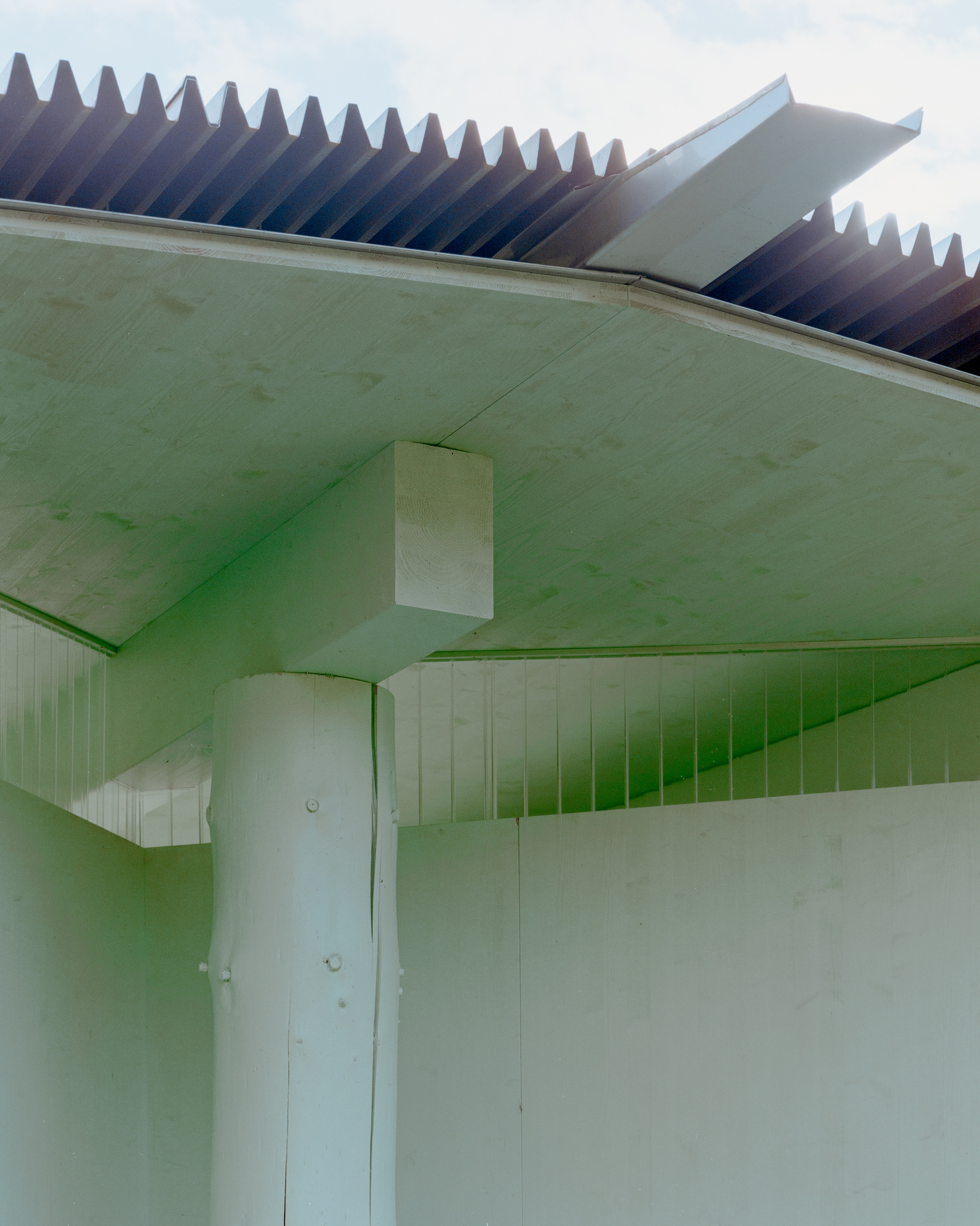 Underside of the pale roof overhang with a round log column, a boxy timber bracket and corrugated roofing against a cloudy sky