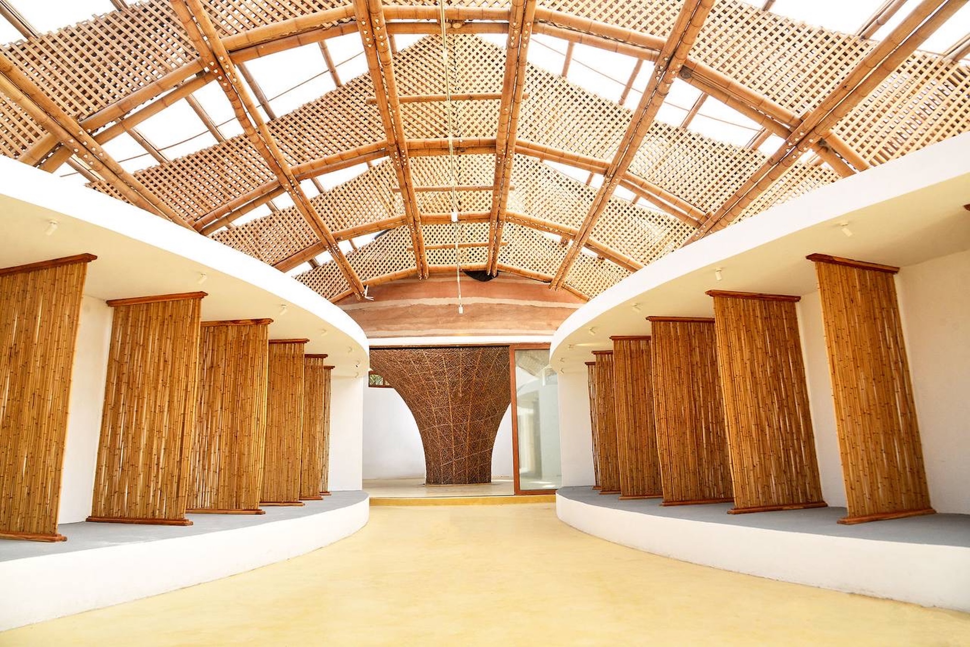 Interior hall with bamboo-clad columns along curved white walls beneath a lattice bamboo vault, daylight filtering through the roof