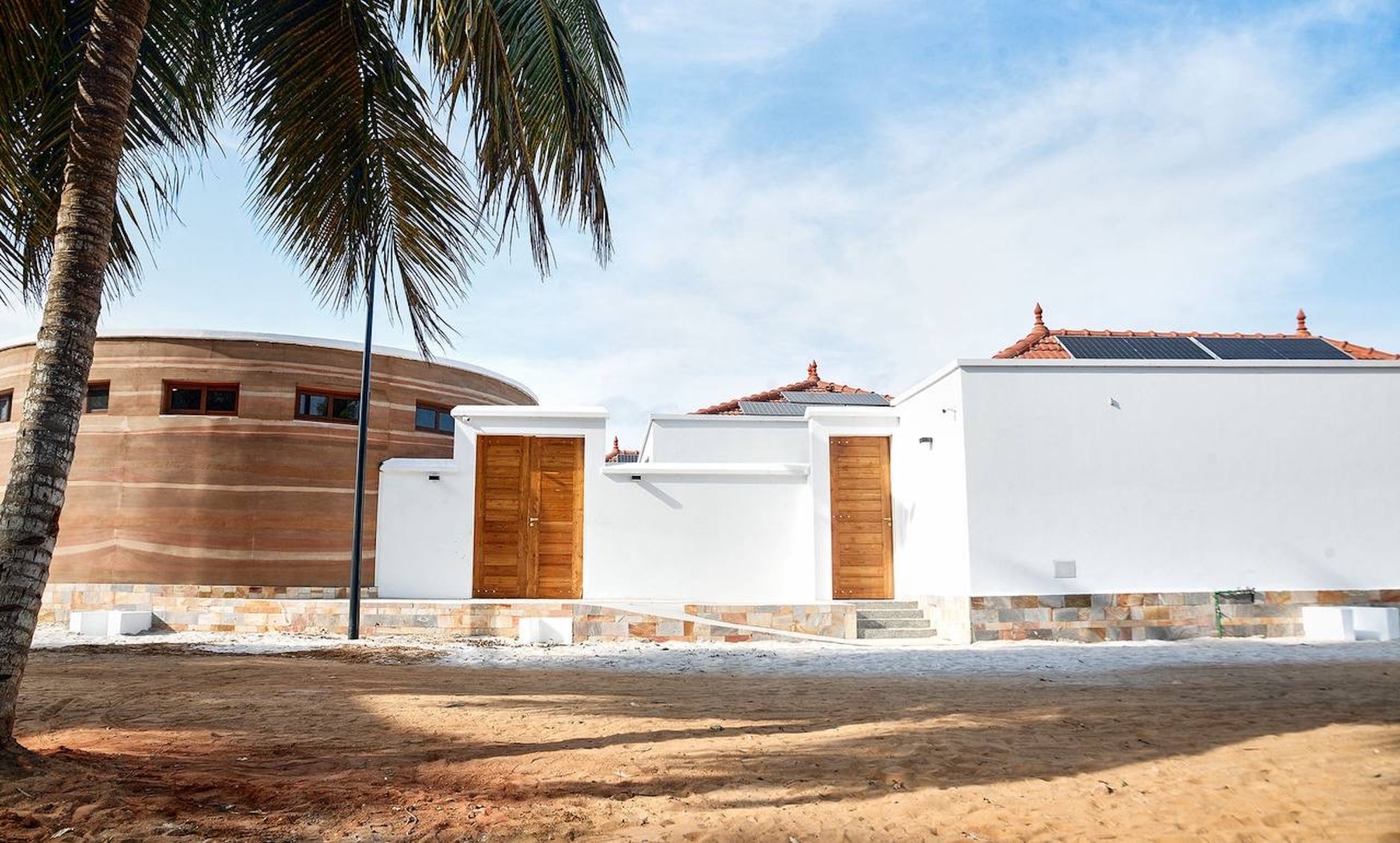 White walls with timber doors beside a round rammed earth volume, seen from sandy ground under a palm frond