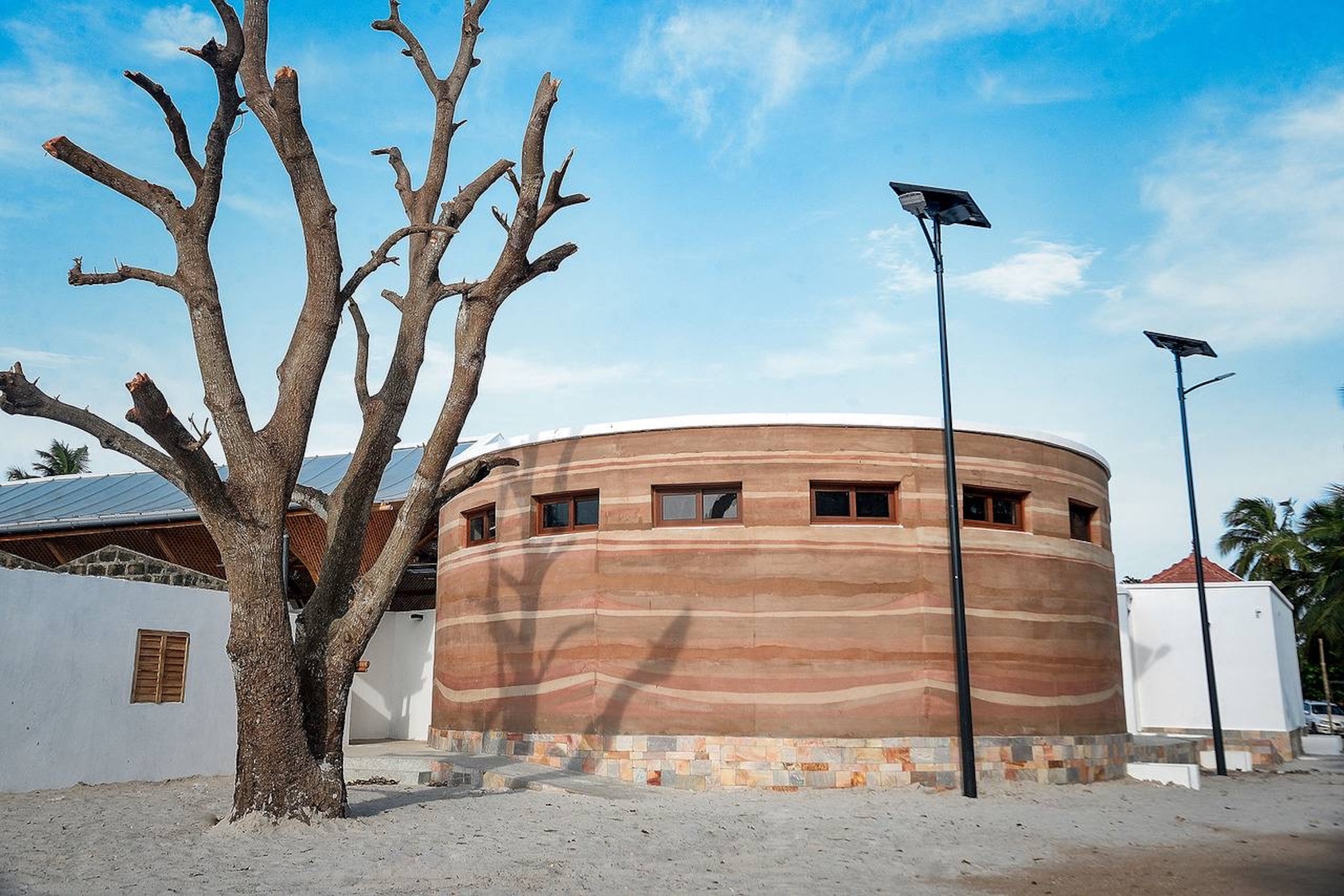 Cylindrical rammed earth building with banded strata and small windows behind a bare tree, solar lamp posts against a blue sky