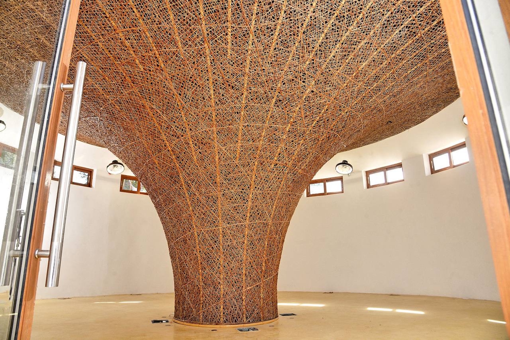 Woven cane column flaring into a funnel-shaped ceiling in a round white room, with small windows and floor reflections of daylight
