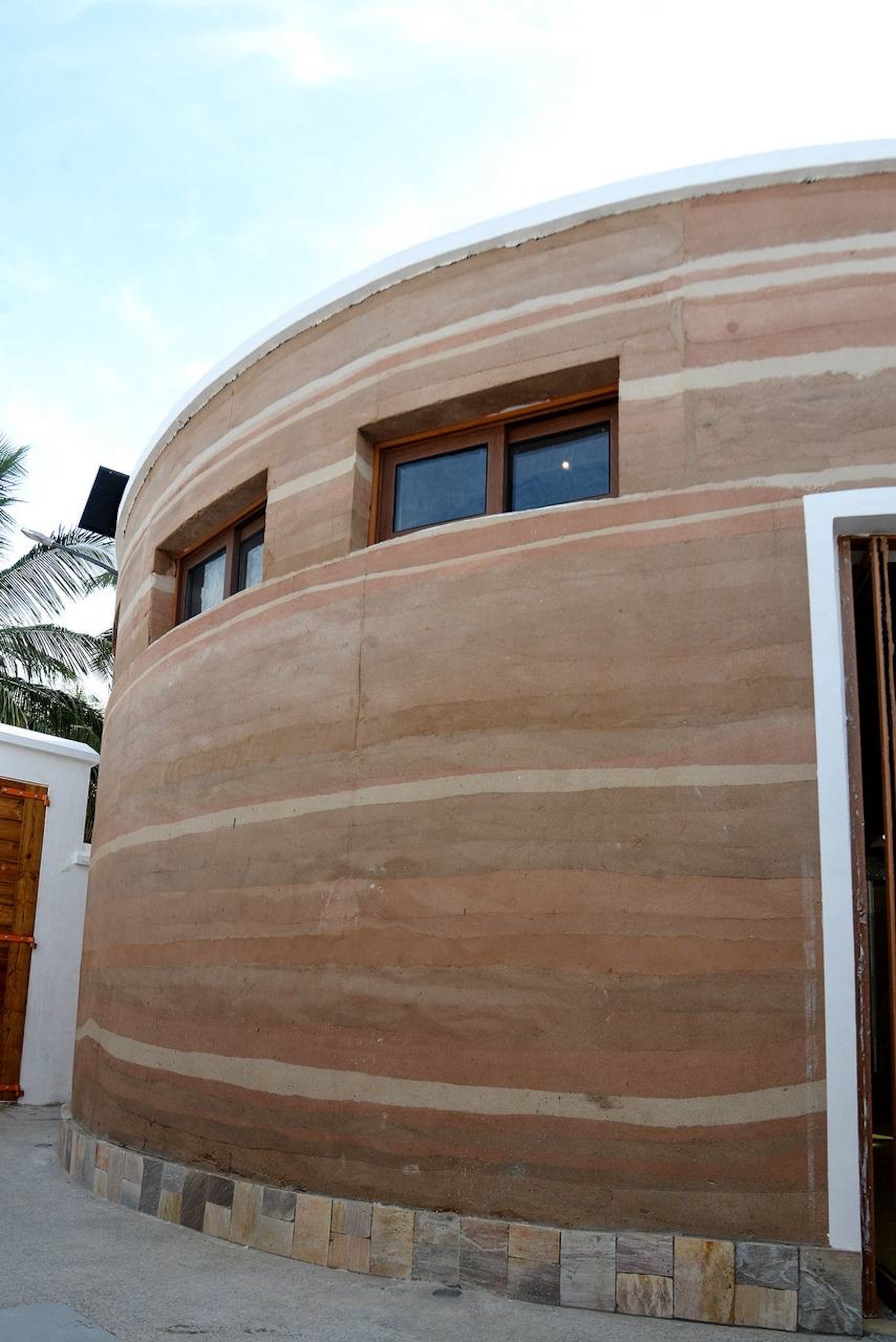 Close view of the curved rammed earth wall with pale and pink layers, timber-framed windows and a stone tile base in bright sun