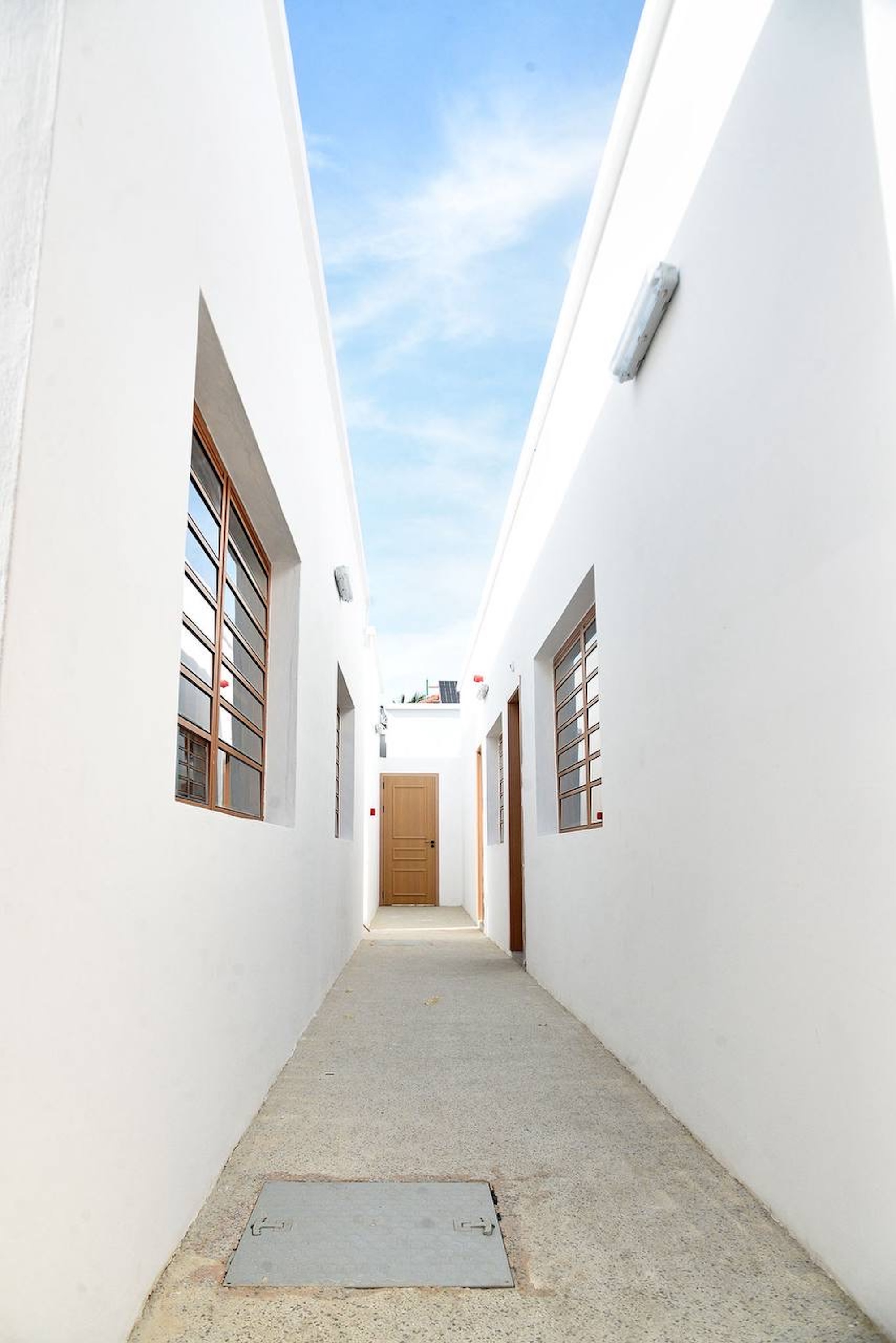 Narrow white-walled passage with timber-framed louvred windows leading to a wooden door under a pale blue sky