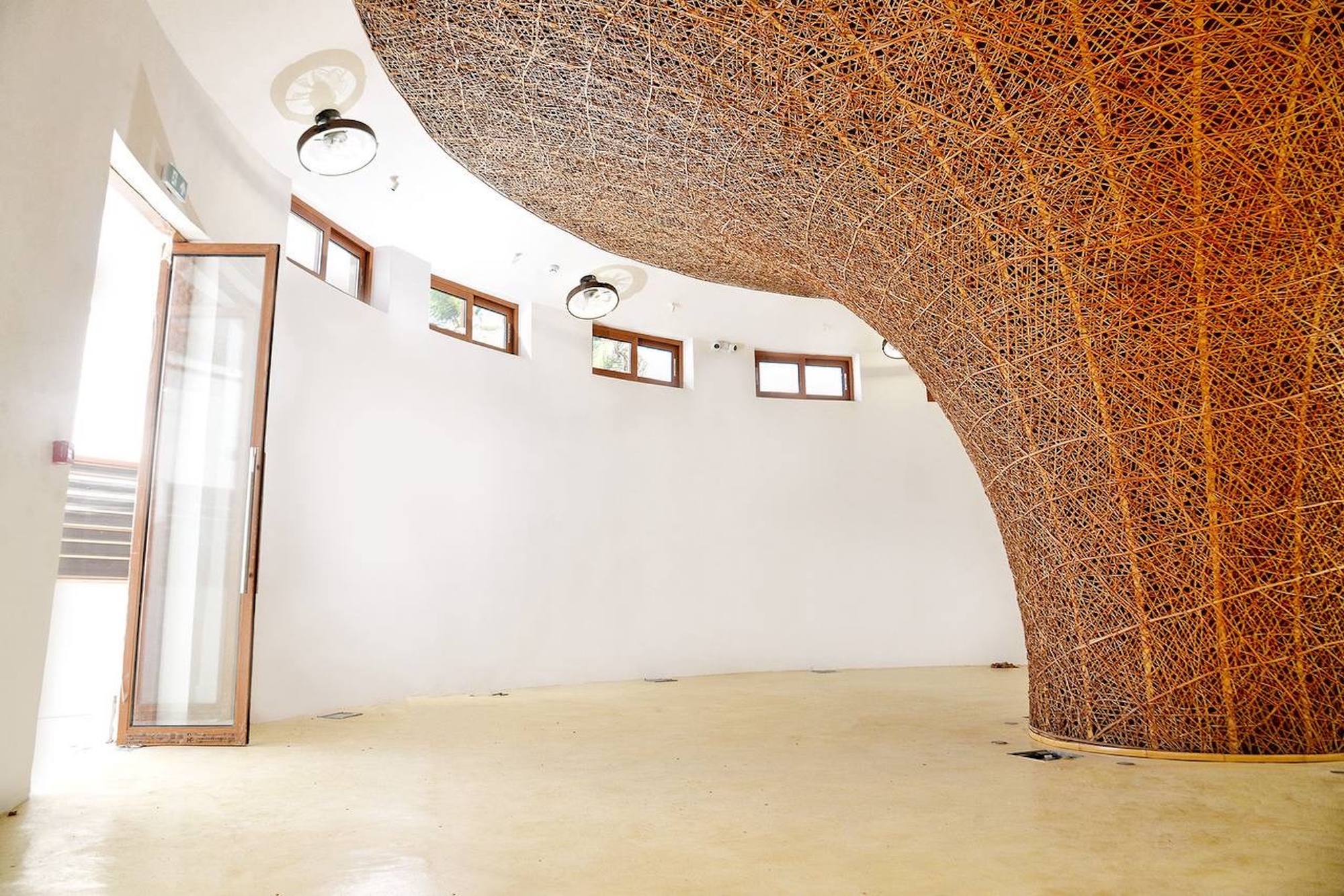 Interior with a curving woven rattan vault sweeping down to the floor beside a glazed timber door in bright daylight