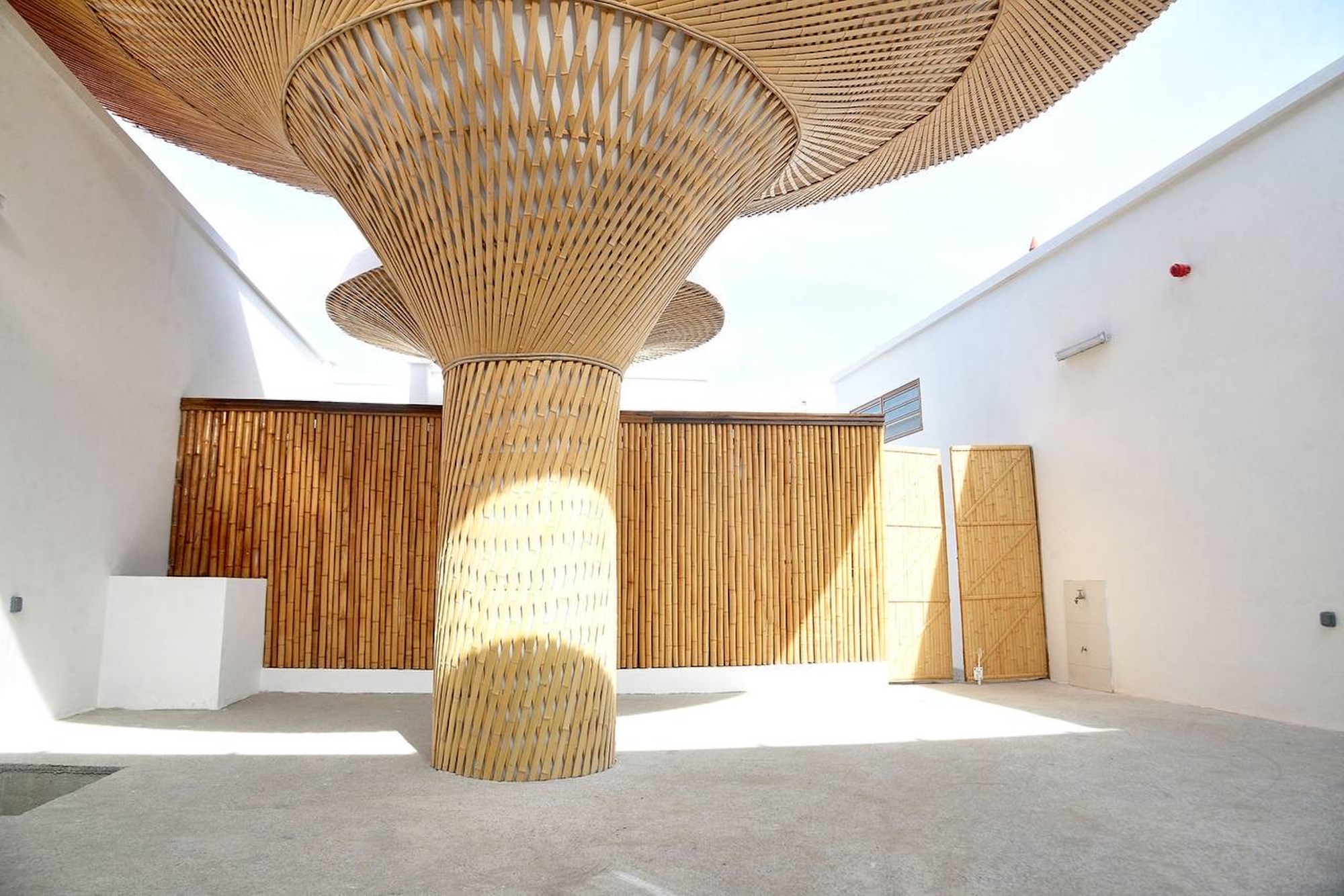Courtyard with a woven bamboo mushroom-shaped column and canopy, bamboo screen wall behind and sunlight cast across the concrete floor