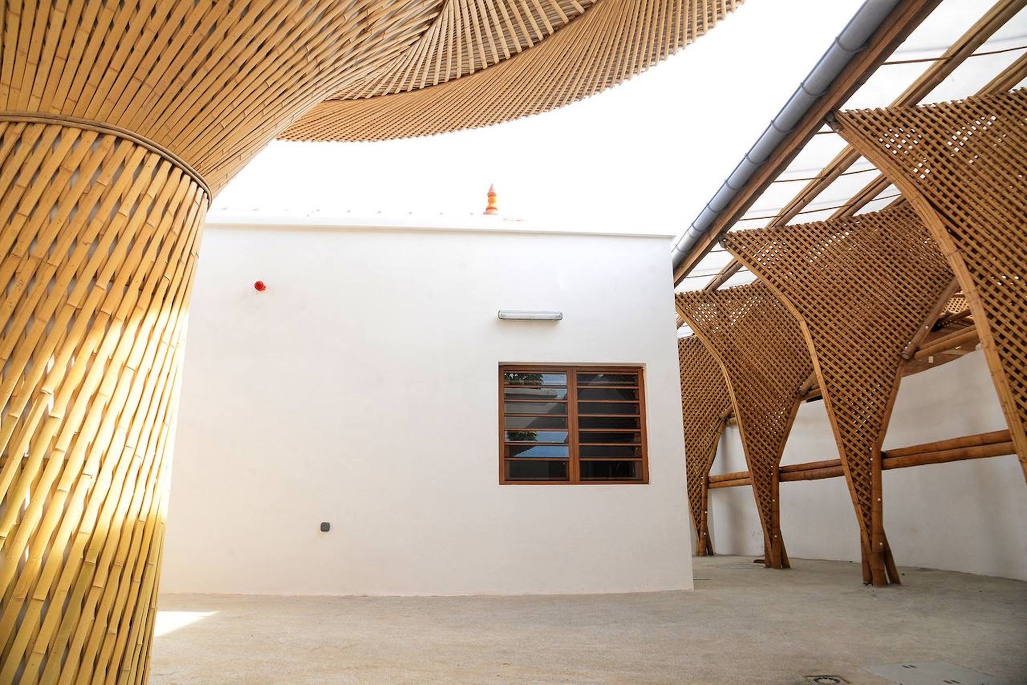 Courtyard view of a bamboo column and canopy beside lattice bamboo arches, with a white wall and timber window under open sky