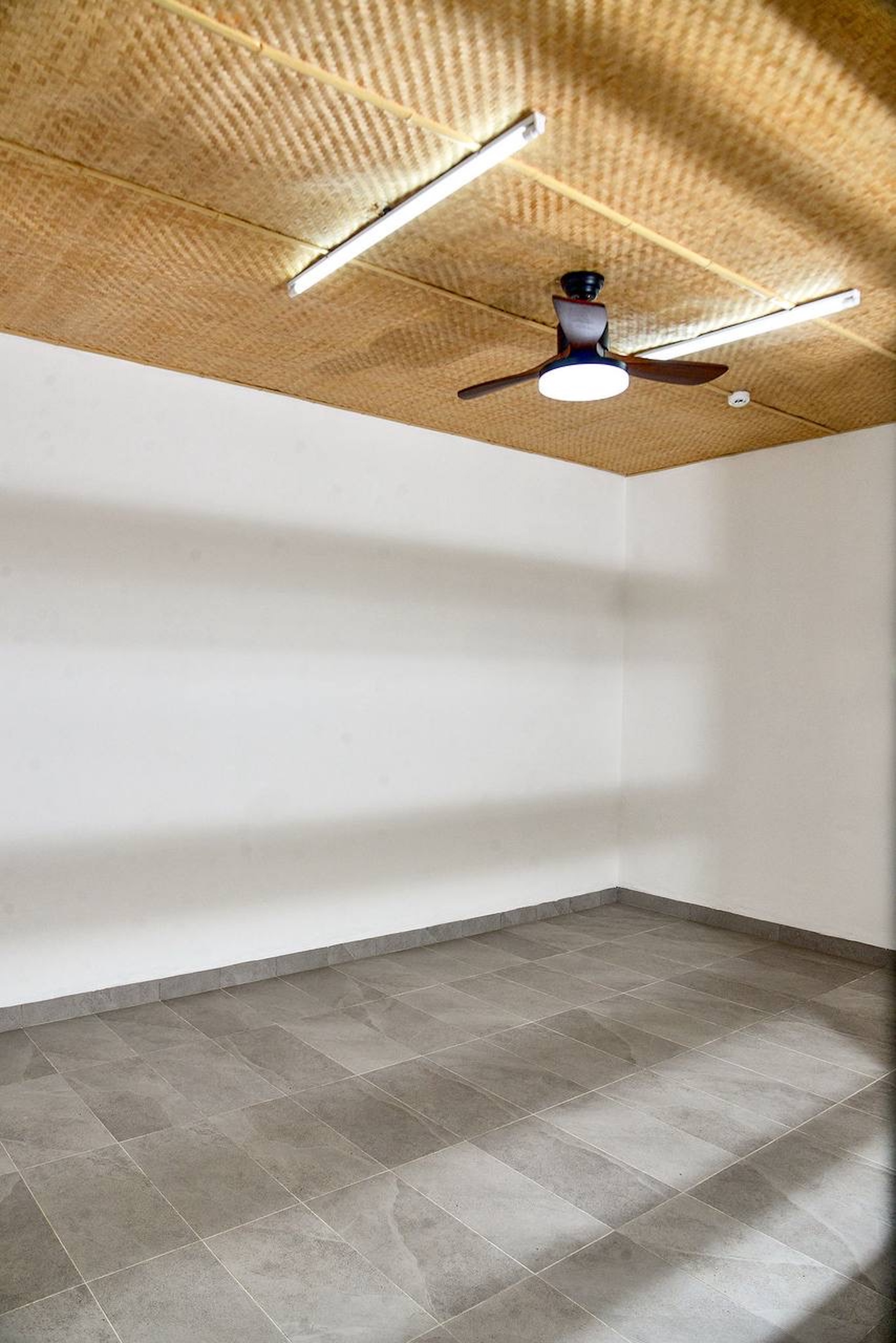 Empty room with a woven bamboo ceiling, strip lights, a dark ceiling fan, white walls and grey stone-look floor tiles