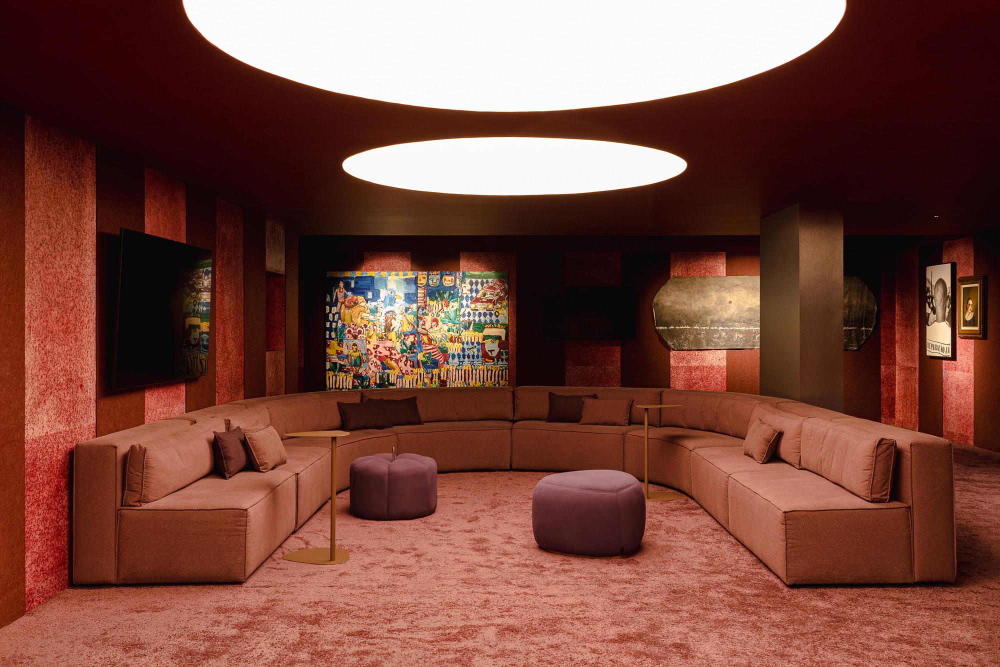 U-shaped sofa in dusty pink facing a colorful painting, with two oval ceiling lights glowing above purple poufs