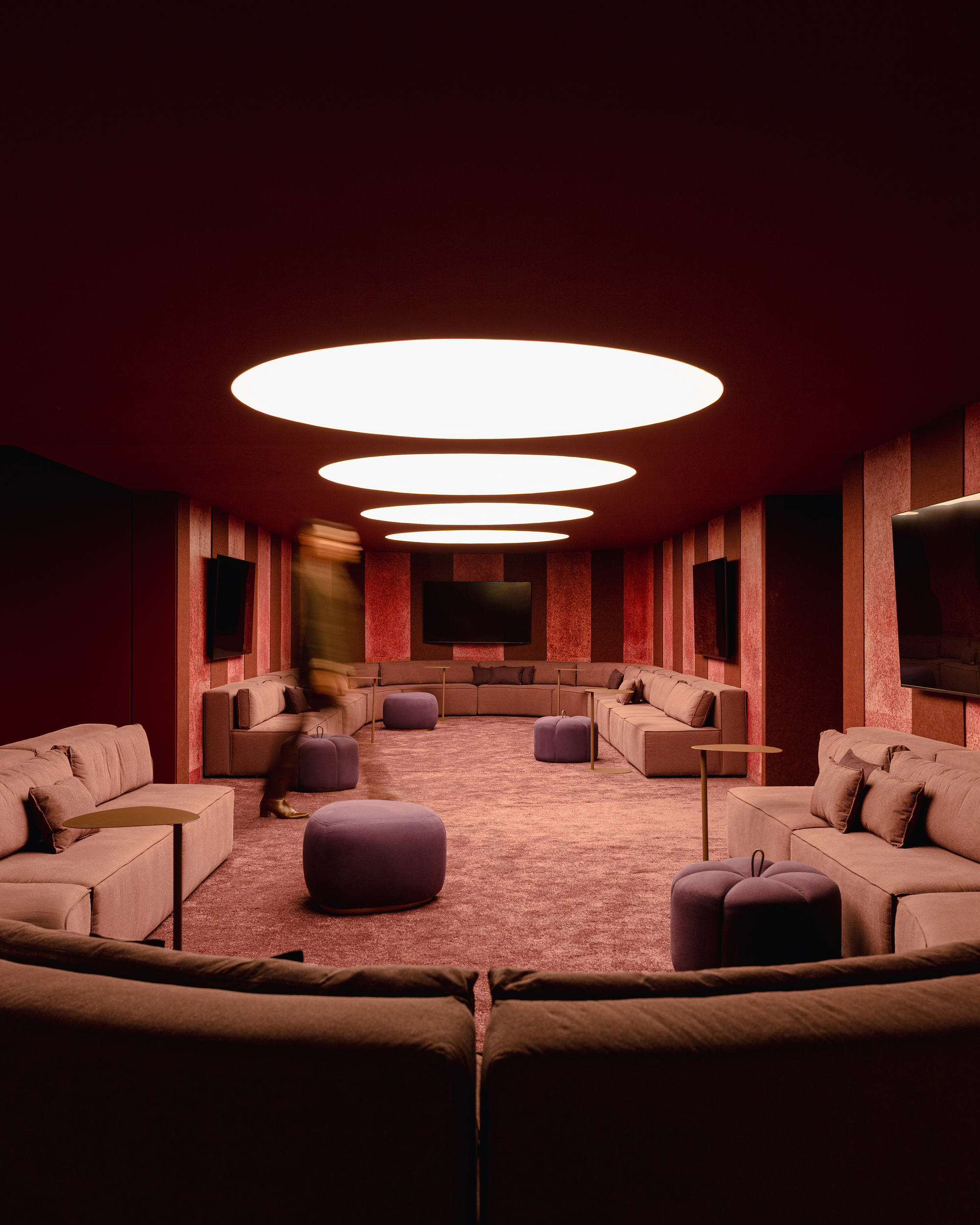 Long lounge with curved sofas and a row of oval ceiling lights, a blurred figure walking past purple poufs