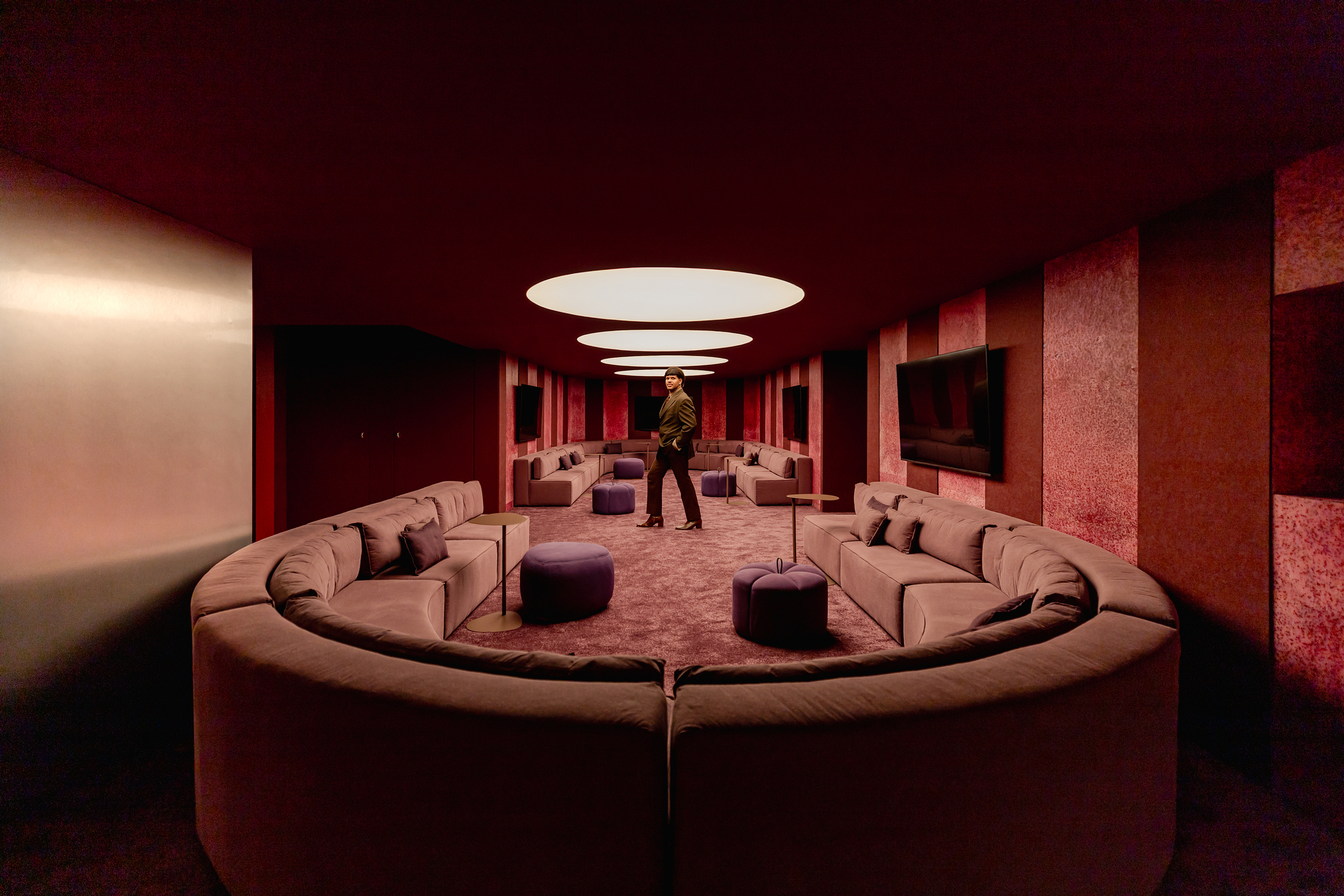 Wide view of the sunken lounge with curved sofas, purple poufs and a standing figure under oval ceiling lights