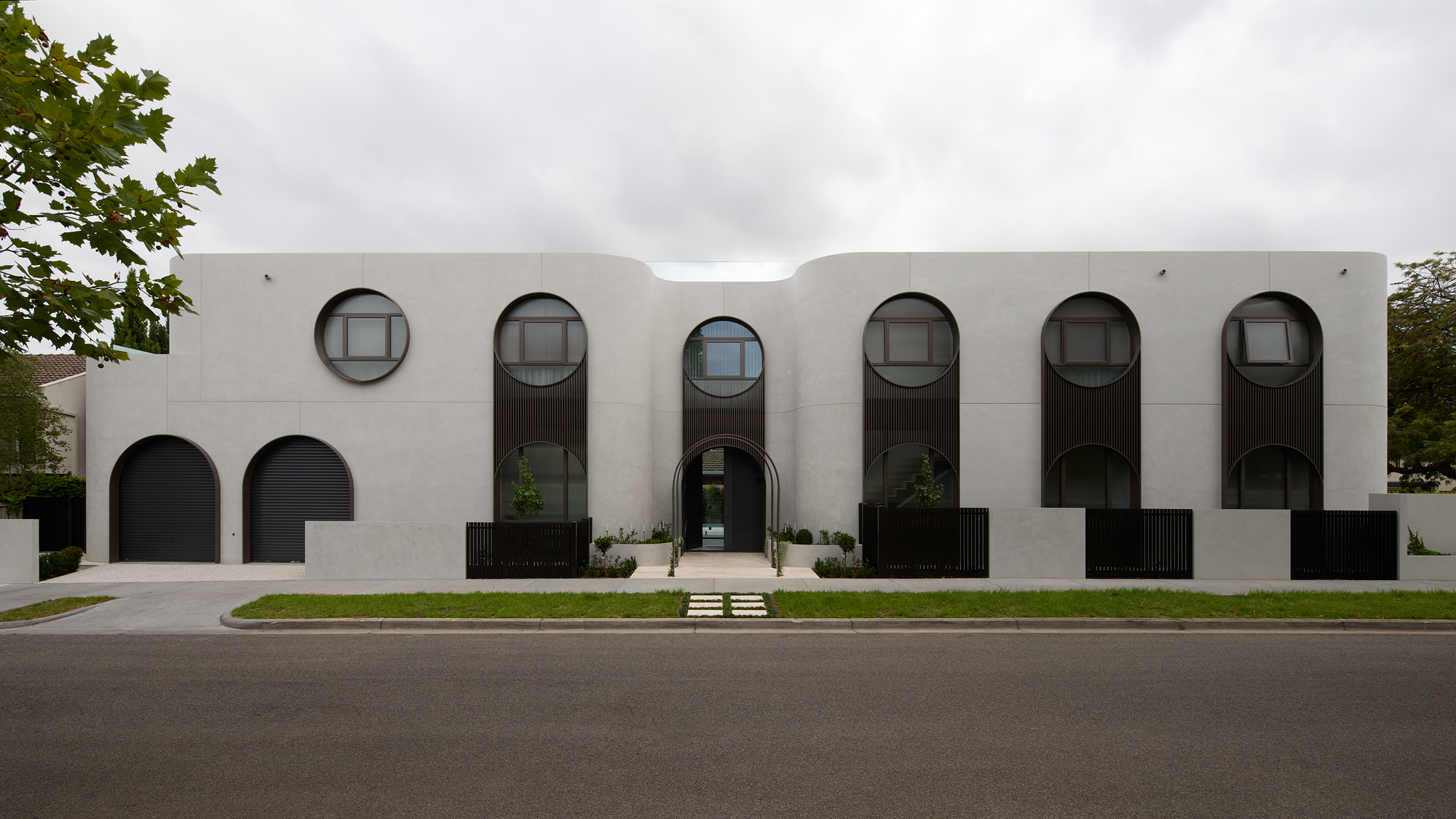 Street facade of pale rendered walls with round and arched dark-framed windows, arched garage doors and a central entry under an overcast sky