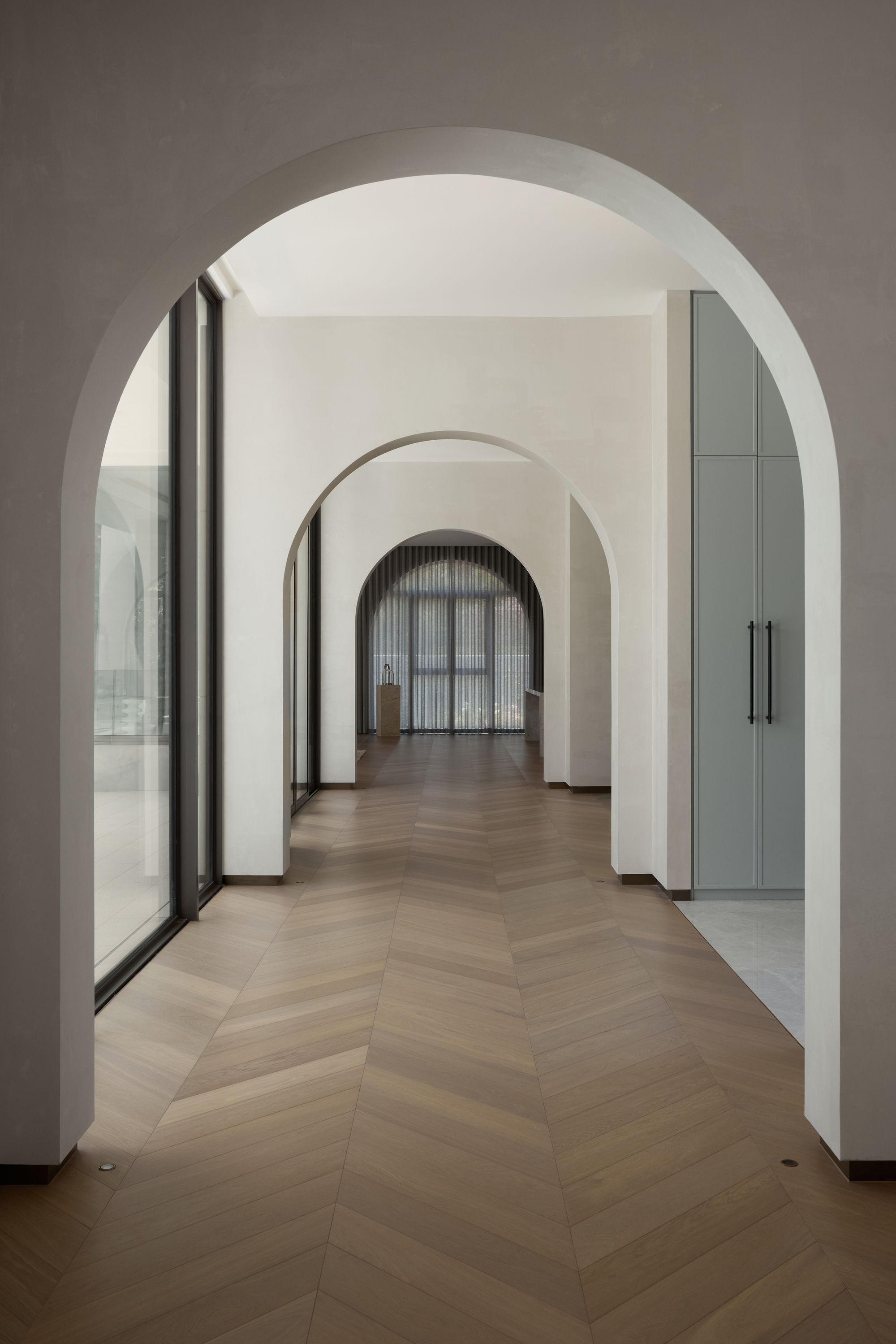 Enfilade of plaster arches along a hallway with chevron timber flooring, a glazed wall on the left and tall cabinetry on the right