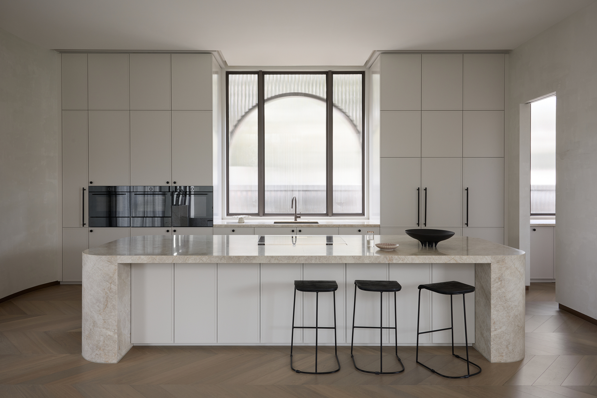Kitchen with a stone-clad island, three black stools and an arched fluted-glass window between tall cabinets, in soft daylight