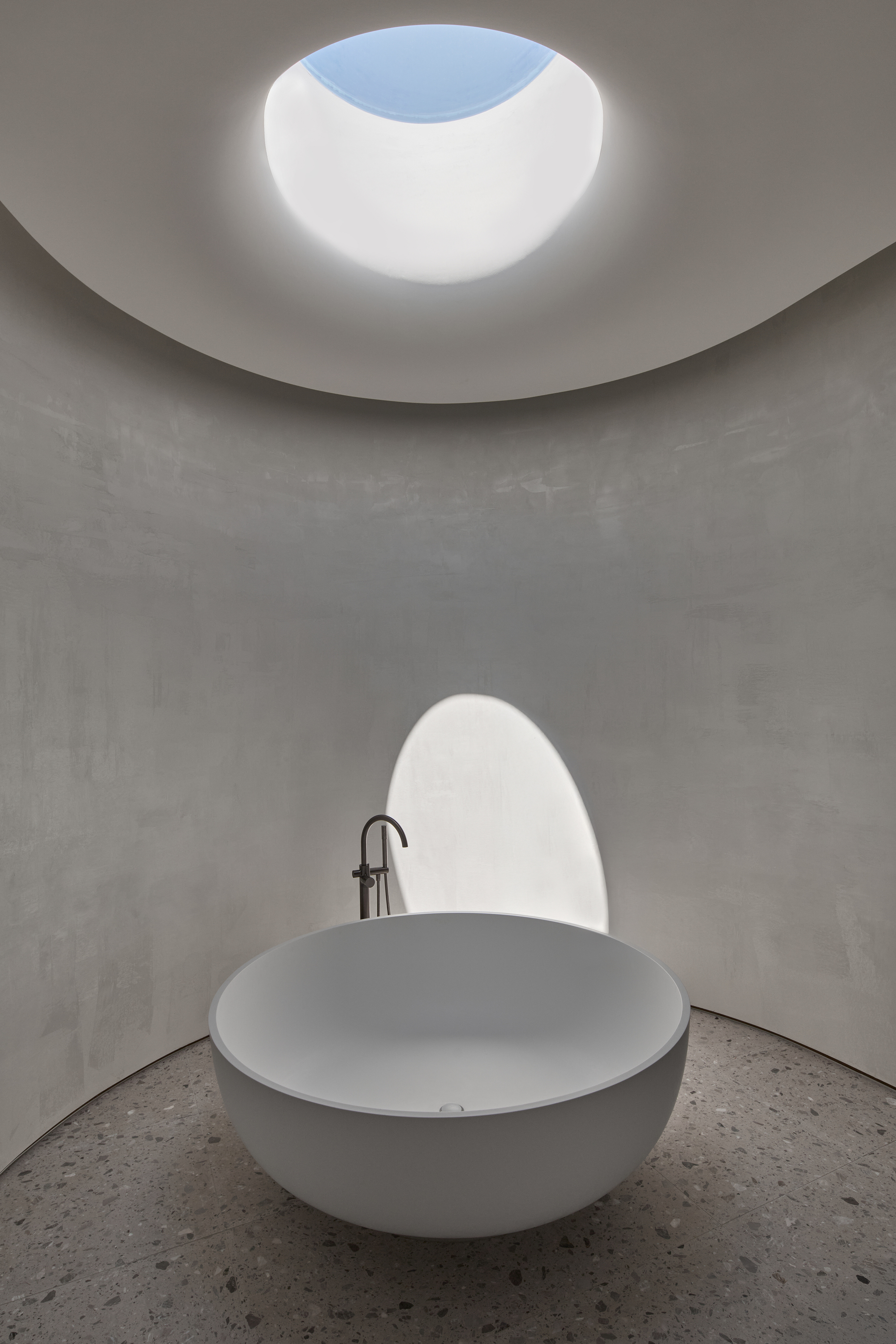 Freestanding round bathtub in a curved plaster room with terrazzo floor, lit by an oval skylight showing blue sky