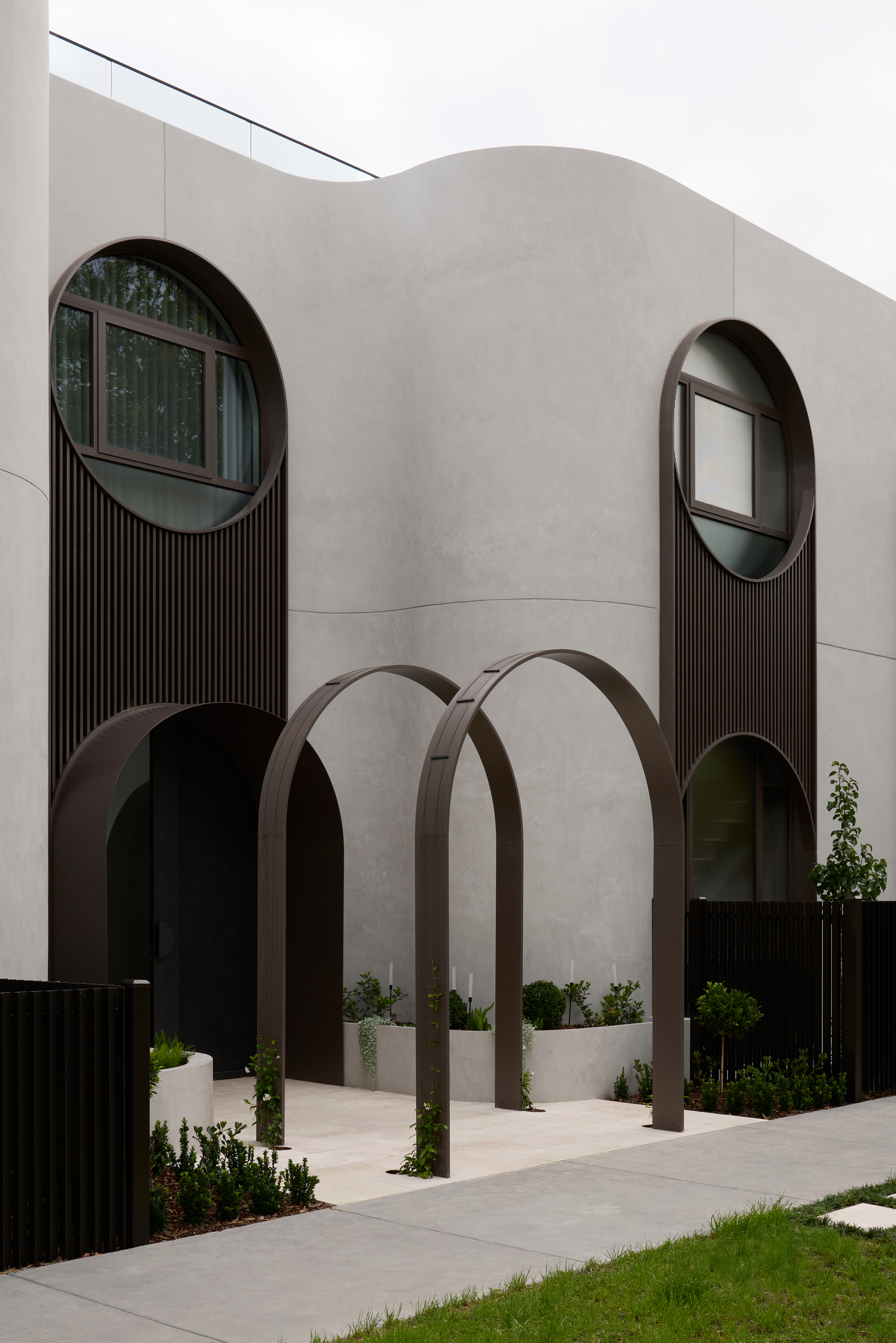 Entry facade with freestanding bronze arches and round windows over vertical timber slats, planted beds in front under overcast sky