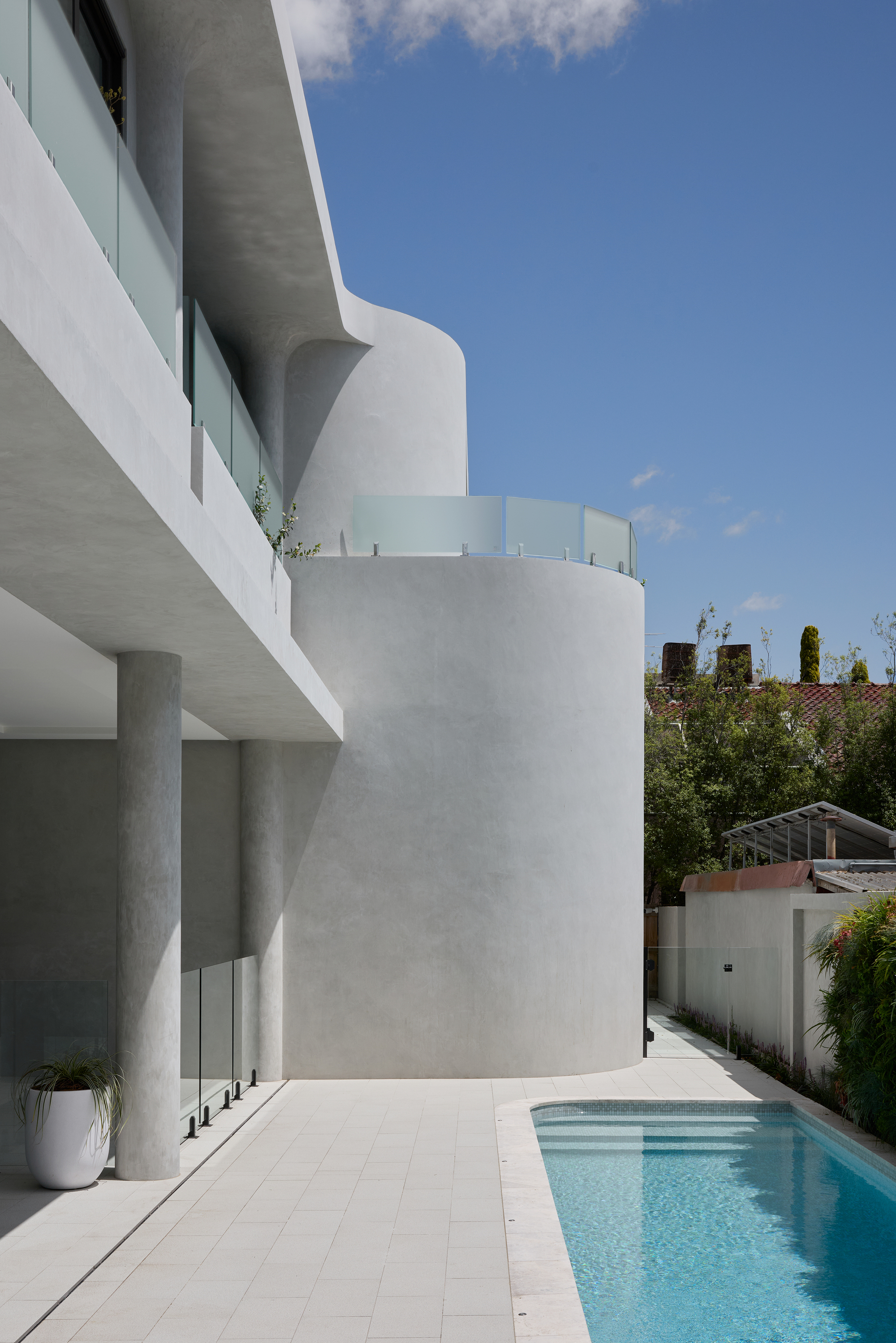 Curved rendered wall and slender round column beside a tiled pool terrace under a clear blue sky