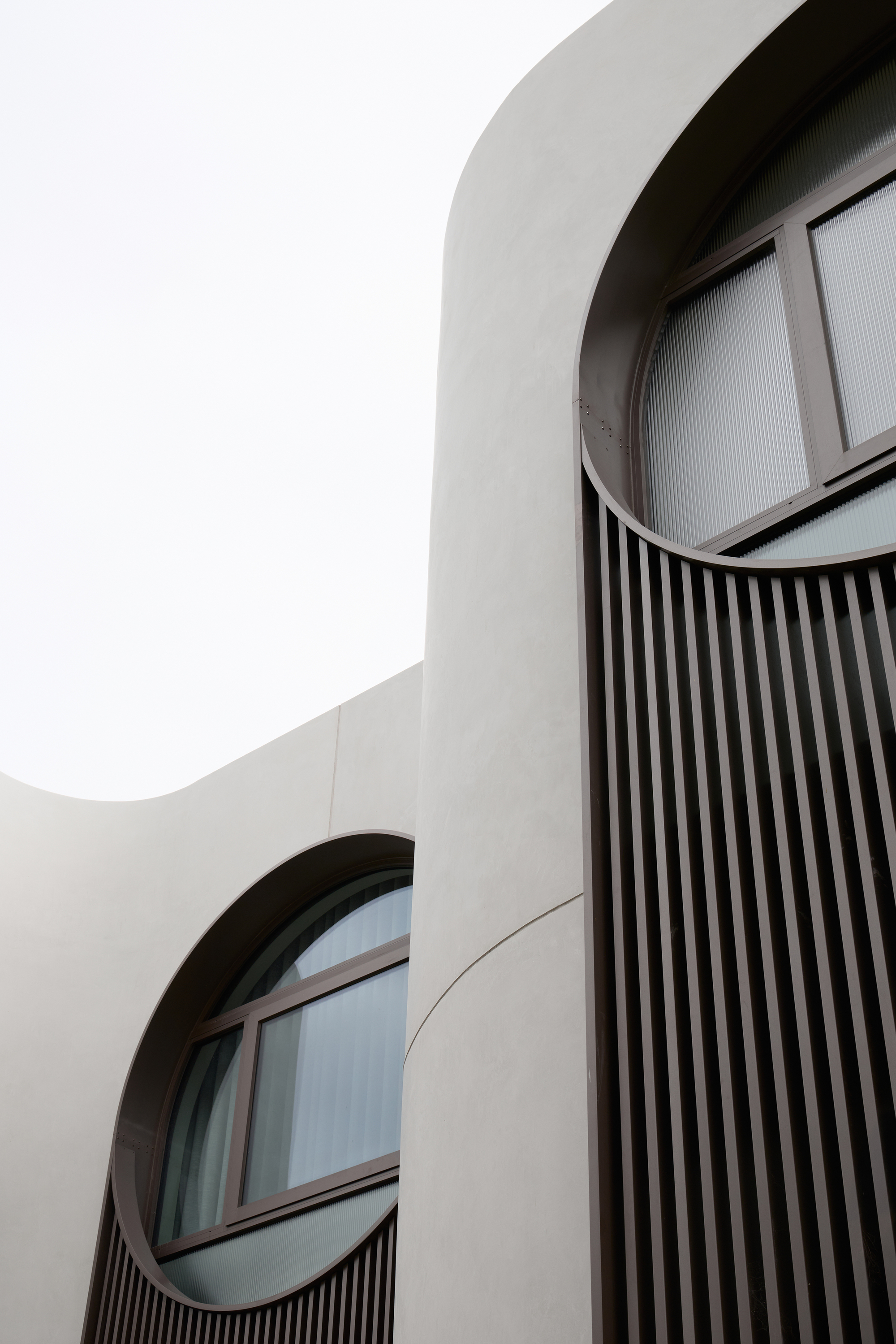Upward view of curved rendered walls with round-topped windows and dark vertical fins against a pale overcast sky