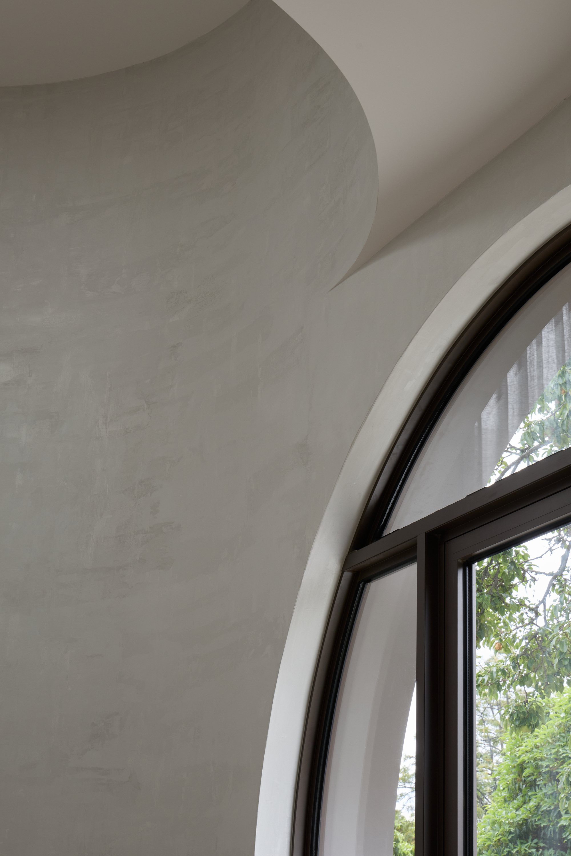 Interior corner of a curved plaster wall meeting an arched dark-framed window, with leafy trees visible outside