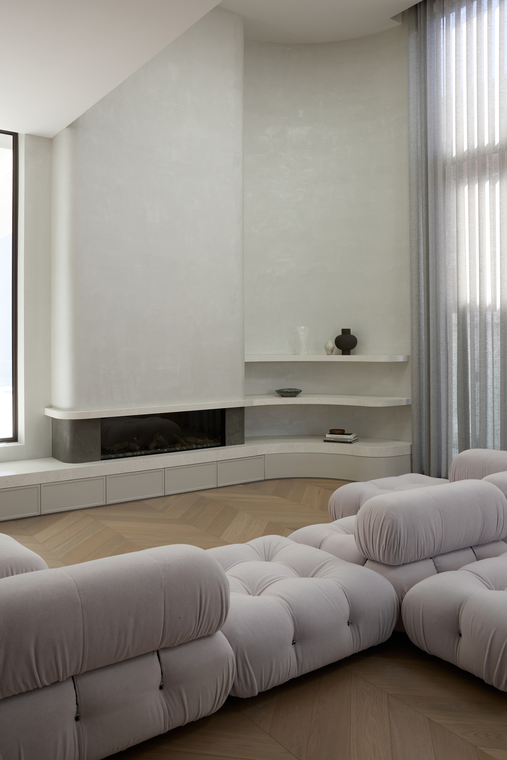 Living room with a low linear fireplace, curved built-in shelf and tufted modular sofa on a herringbone floor beside sheer curtains