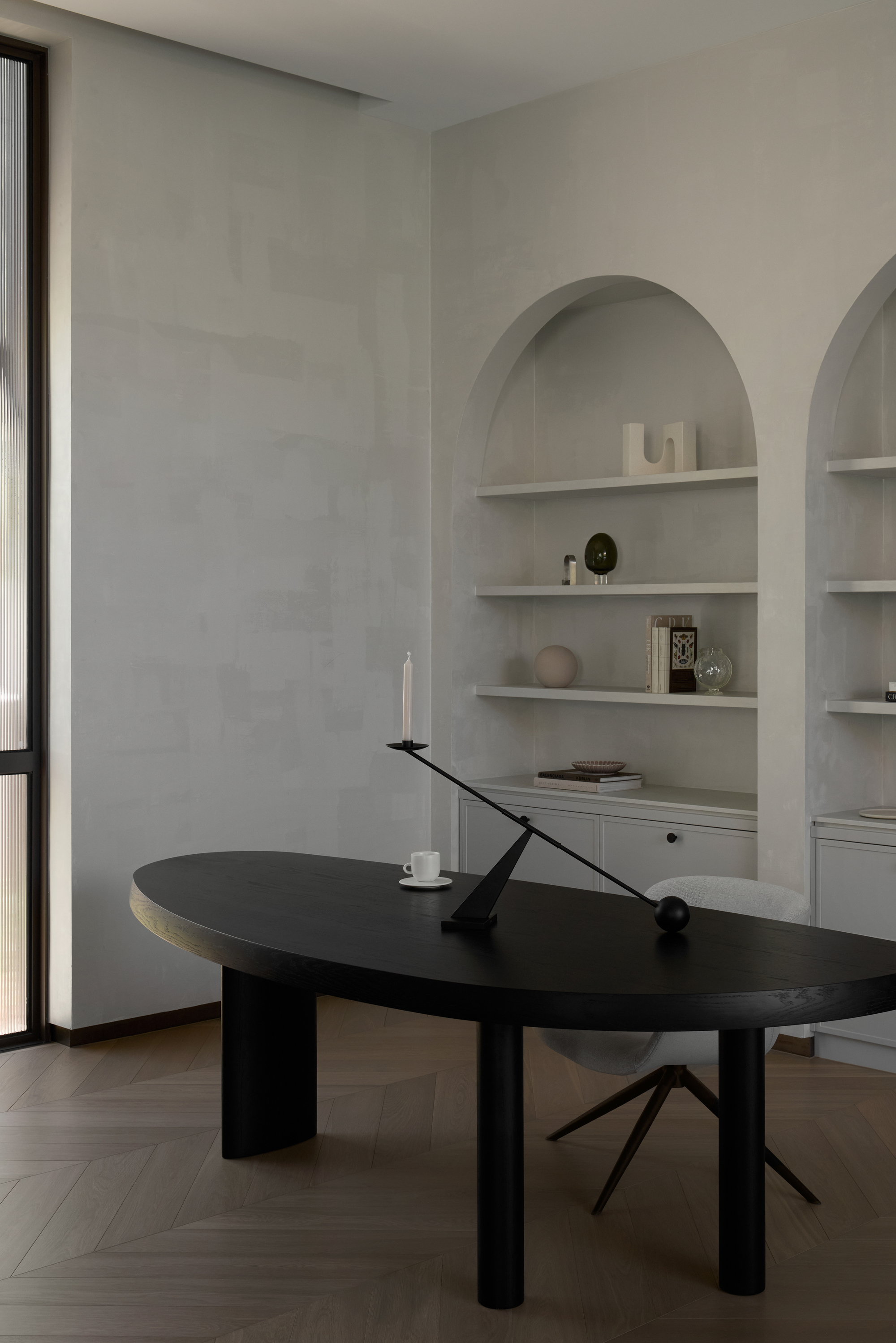 Black oval desk with a counterbalance candleholder in front of arched built-in shelves, with soft daylight from a tall window