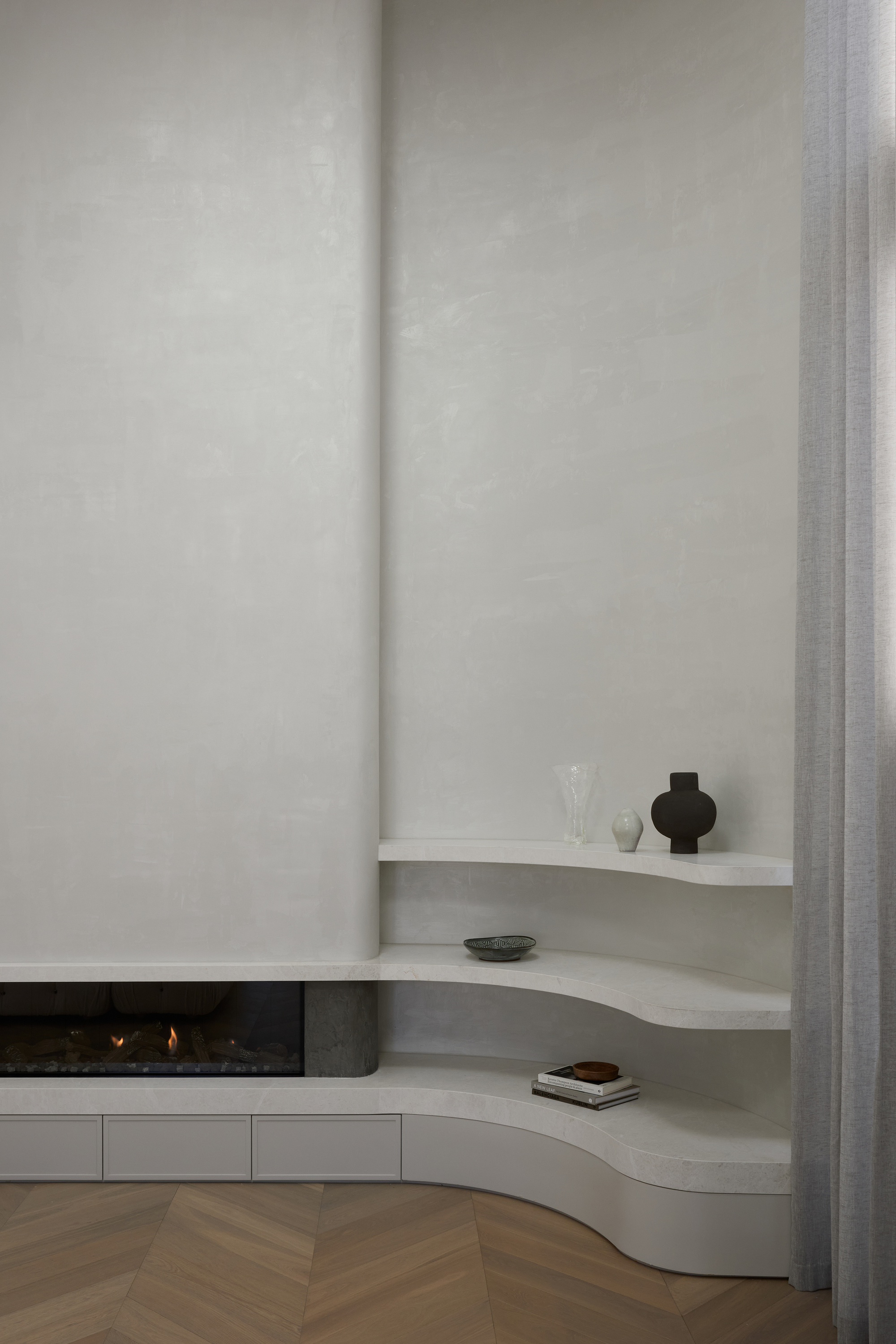 Curved plaster fireplace wall with stepped stone shelves, a low gas flame and a sheer curtain at the edge
