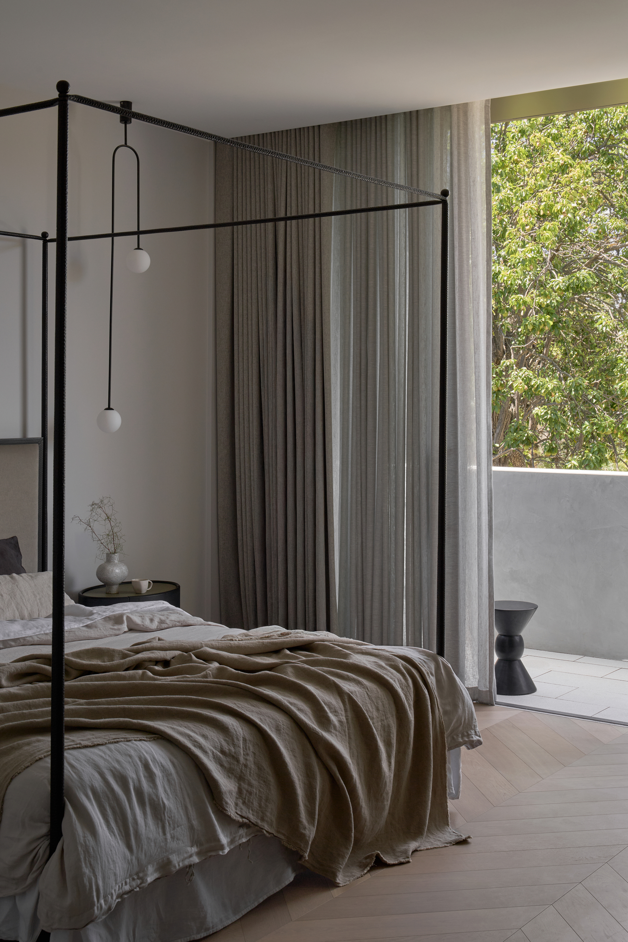 Black four-poster bed with linen bedding and pendant lights, curtains drawn back to a balcony and leafy tree