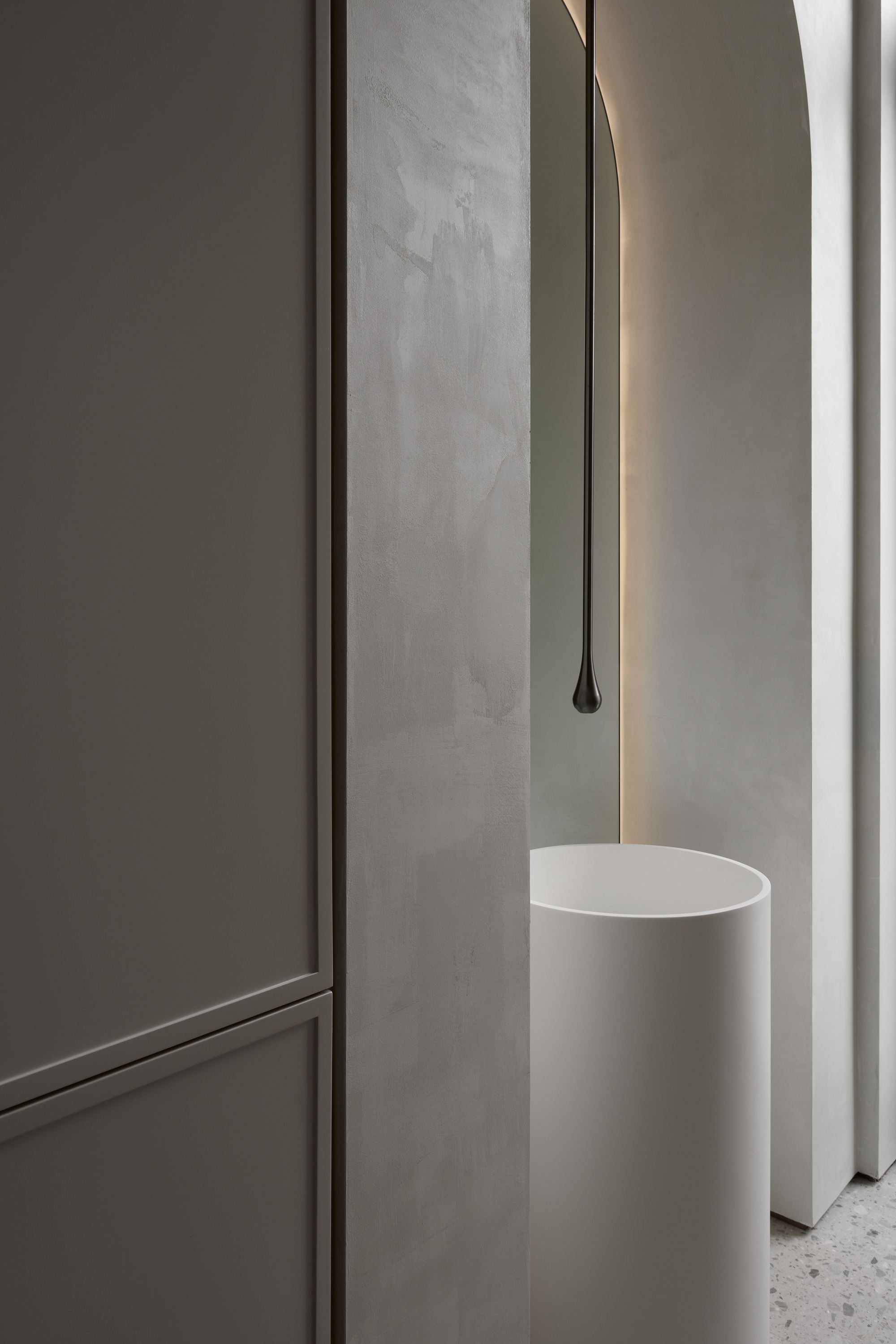 Freestanding white basin beneath a thin droplet-shaped tap, beside a plaster column and an arched niche with warm backlight