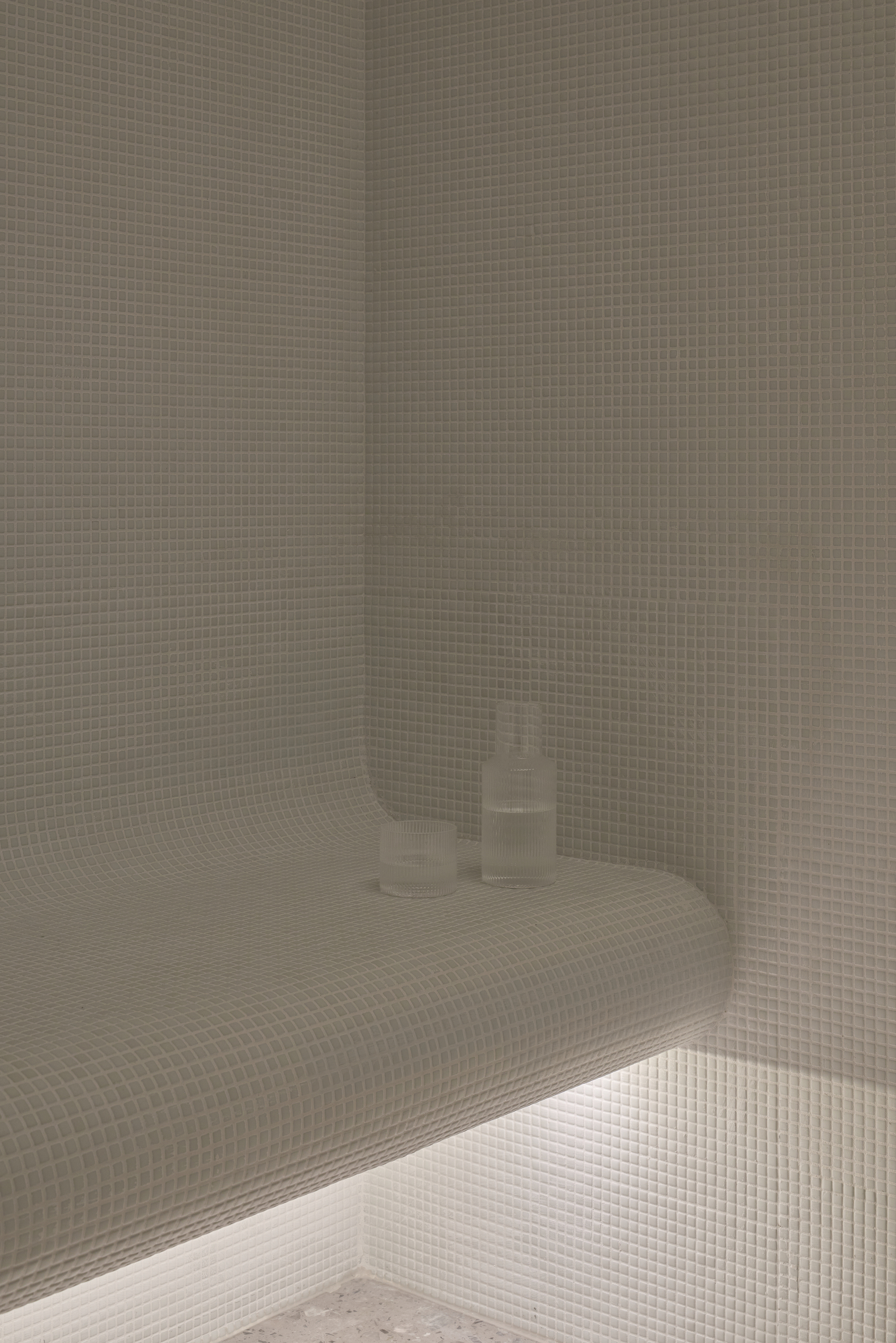 Curved mosaic-tiled steam room bench with a carafe and glass, lit by a glowing strip beneath the seat
