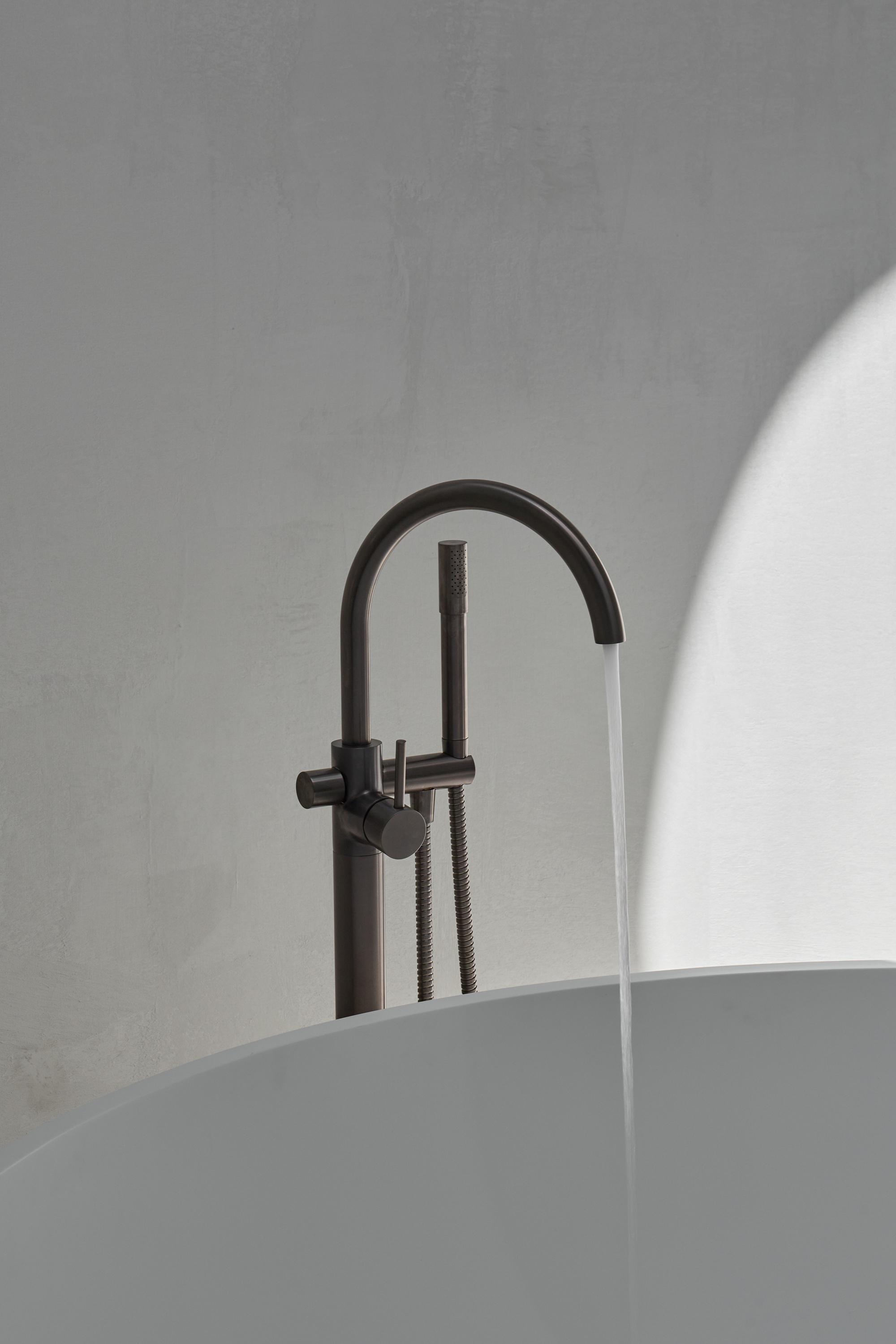 Freestanding bath mixer in dark metal with a handheld spray, water pouring into the tub as a sunlit arch falls on the wall