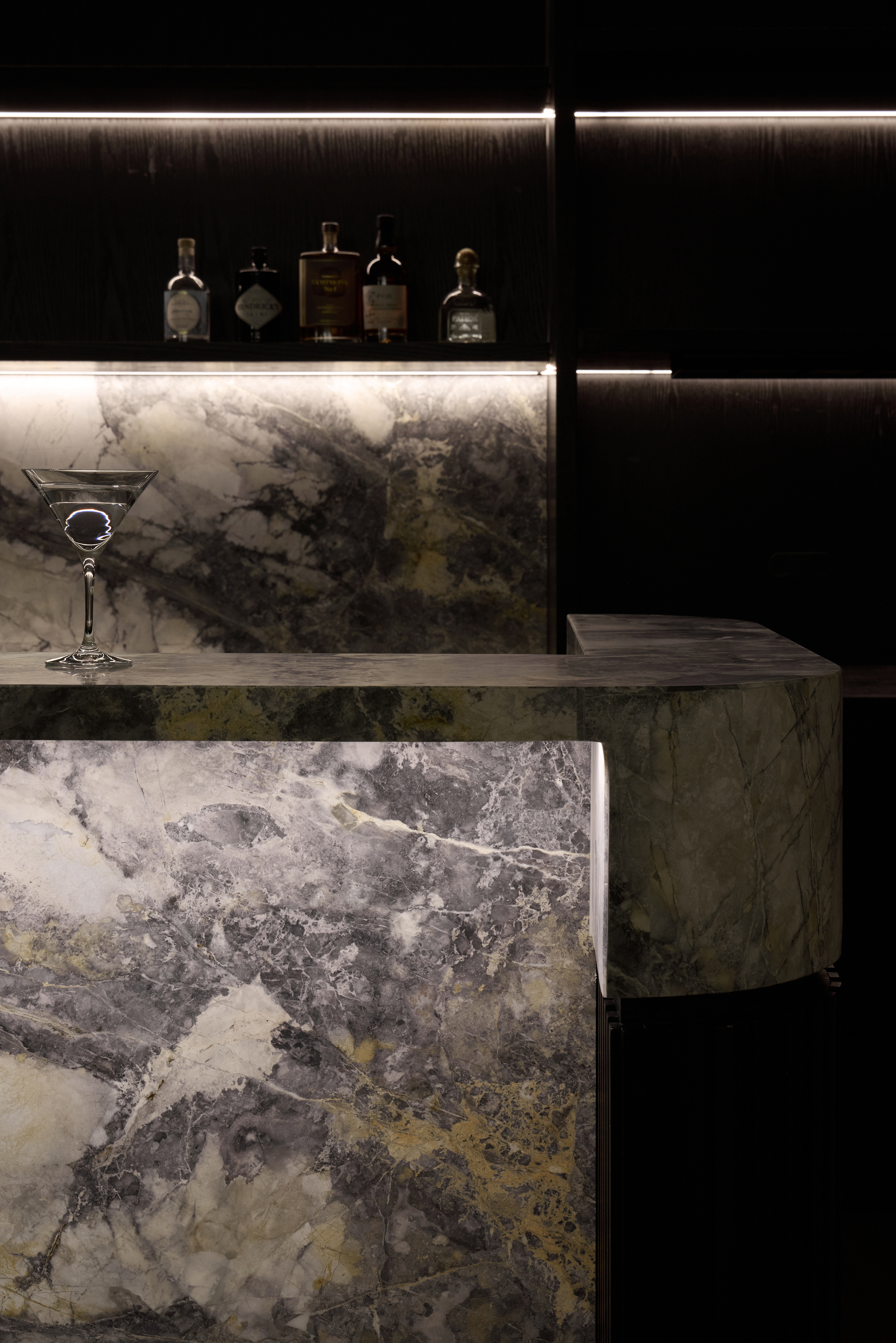 Marble bar with veined green and grey stone, a martini glass on the counter and backlit bottles on the shelf behind