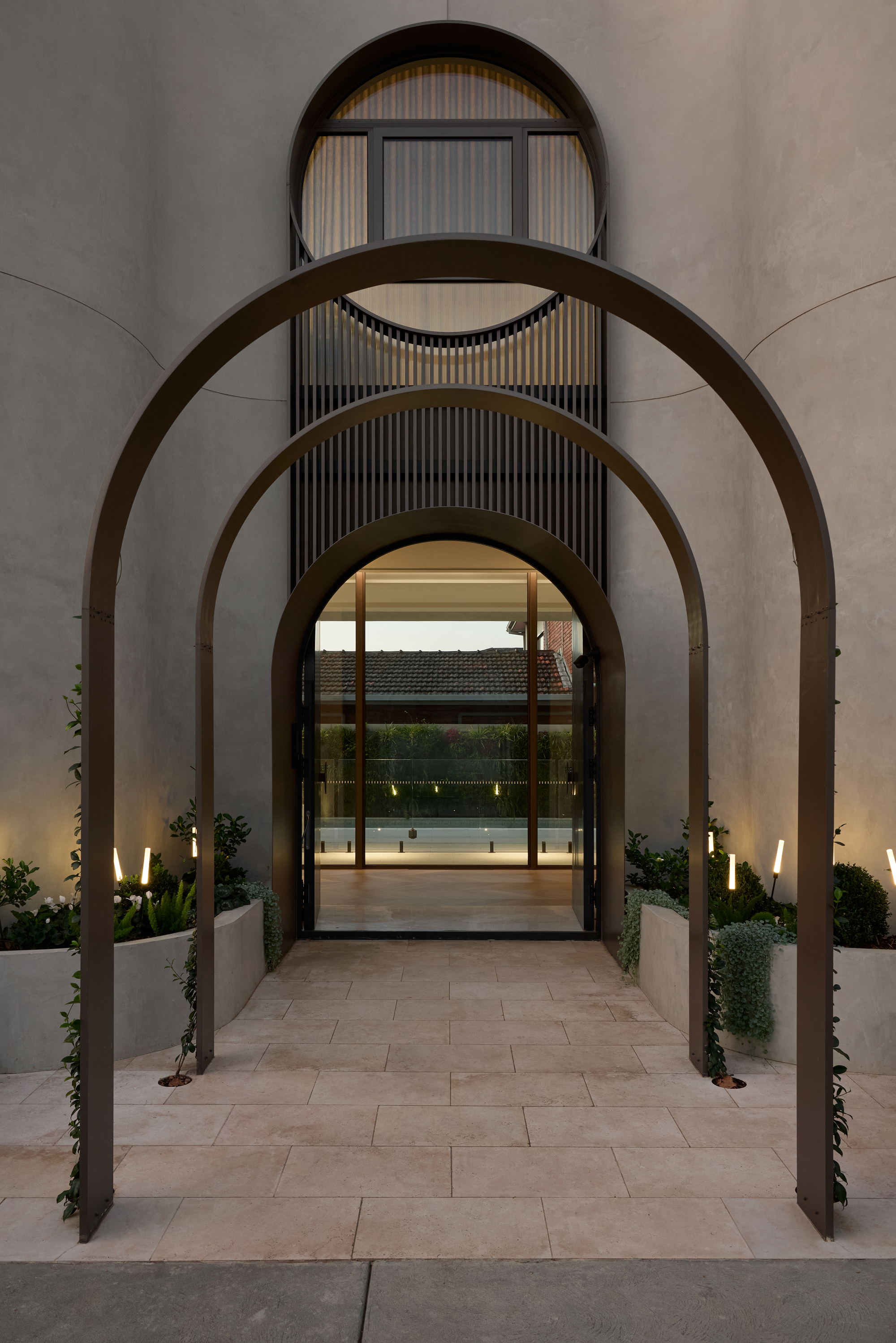 Three bronze arches over a stone-paved entry path, with planted beds and low lights at dusk, and a round window above