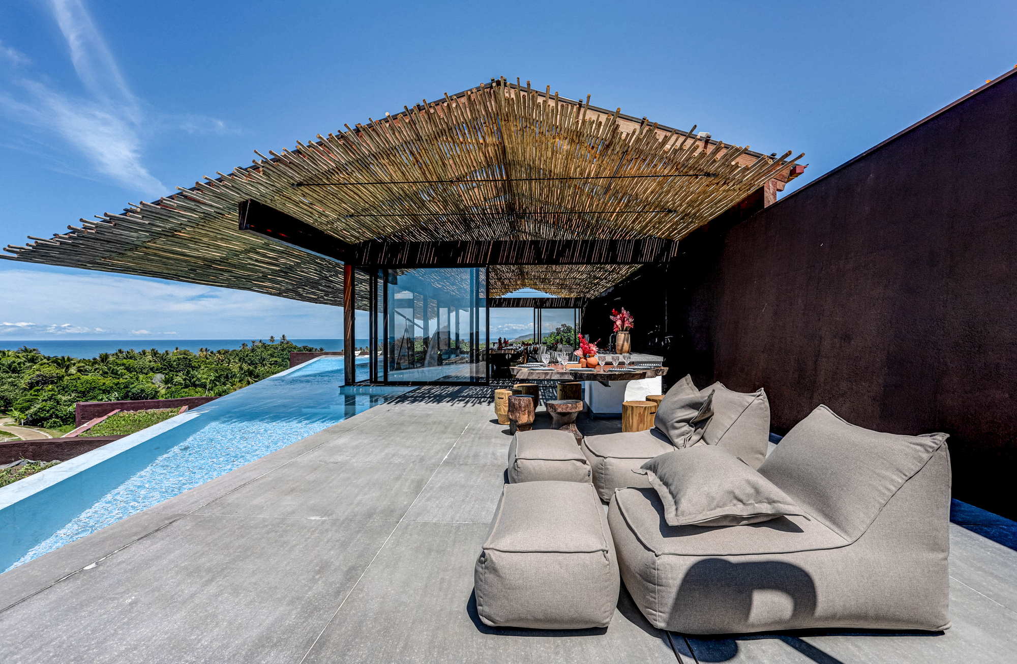Terrace with a woven bamboo canopy over glazed living space, an infinity pool edge and low bean-bag seating under a clear blue sky