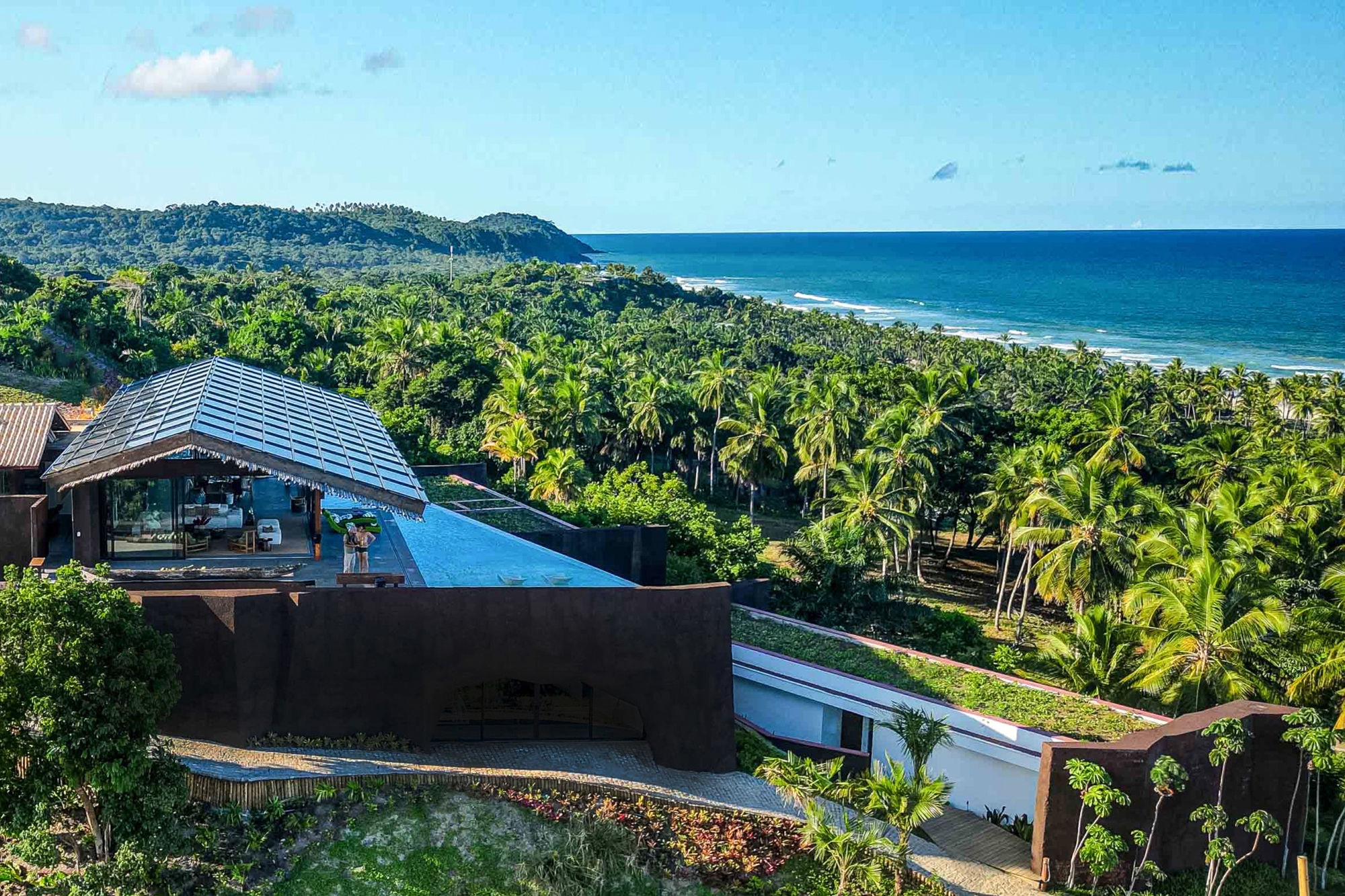 Aerial view of the metal-roofed pavilion and dark rusted walls above a coconut palm grove, with the ocean beyond