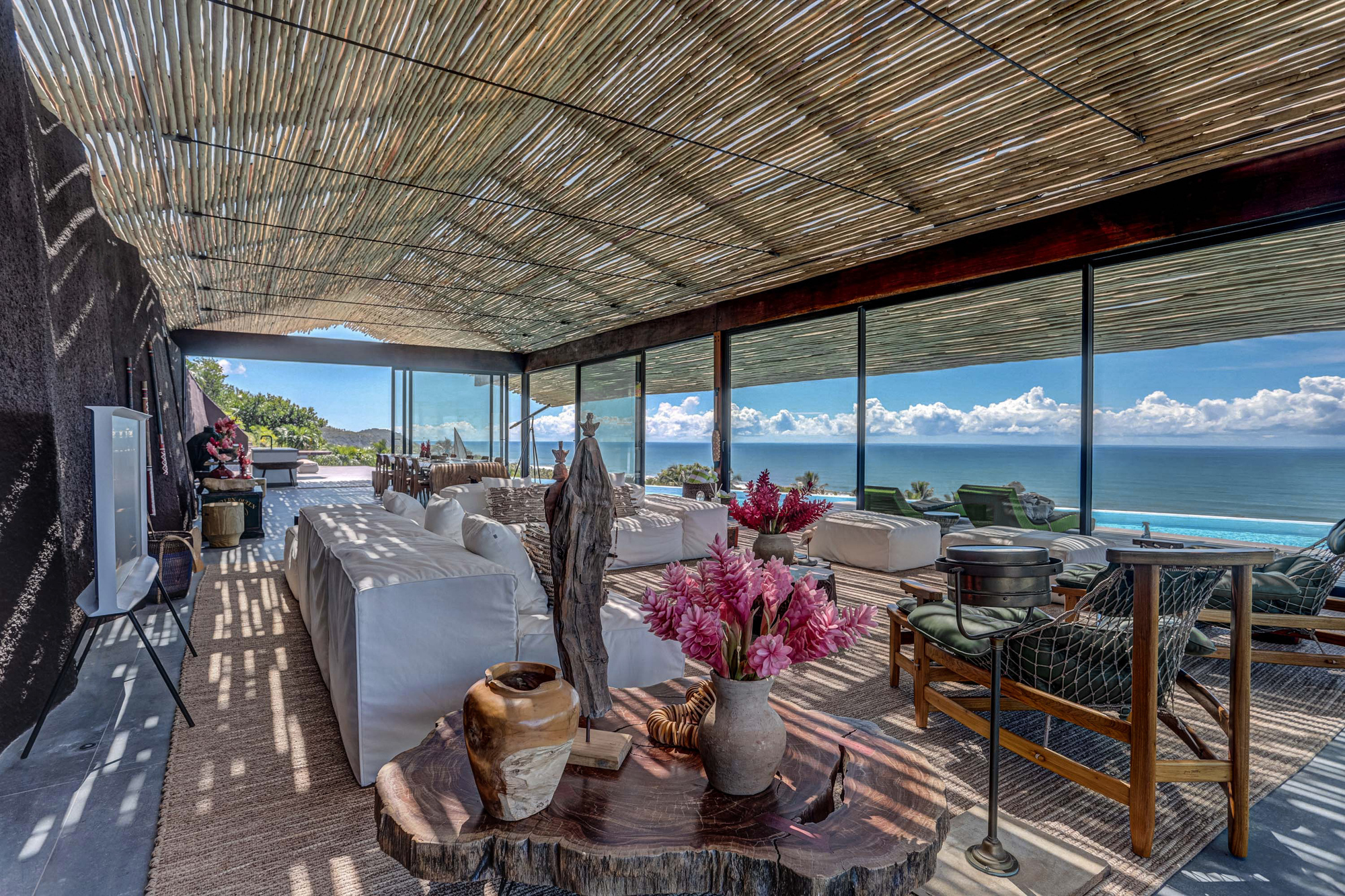 Living area under a slatted cane ceiling with white sofas, pink flowers and sea views, dappled sunlight falling across the rugs