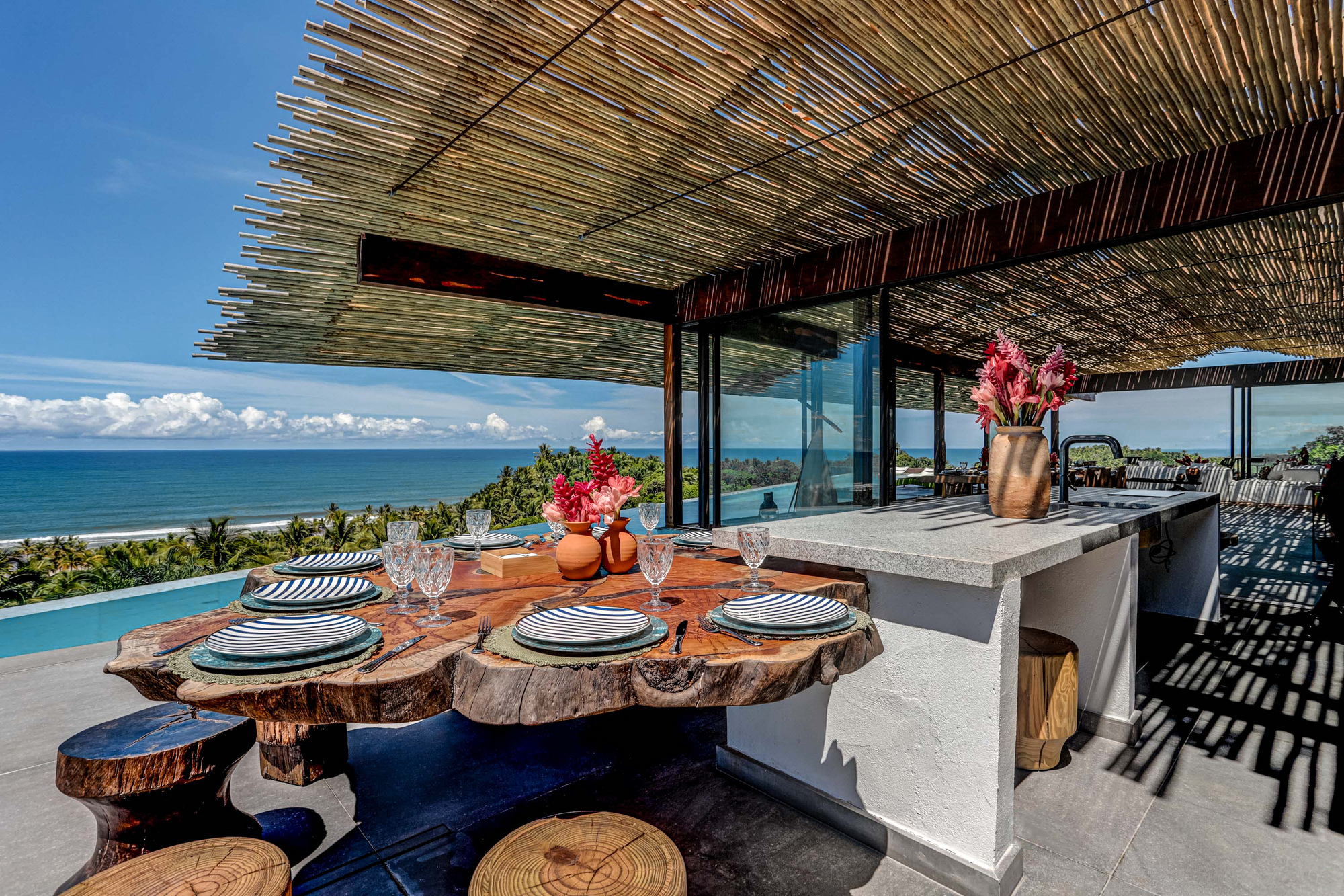 Outdoor dining table made from a live-edge slab beside a white kitchen counter, set under the cane roof with ocean horizon behind