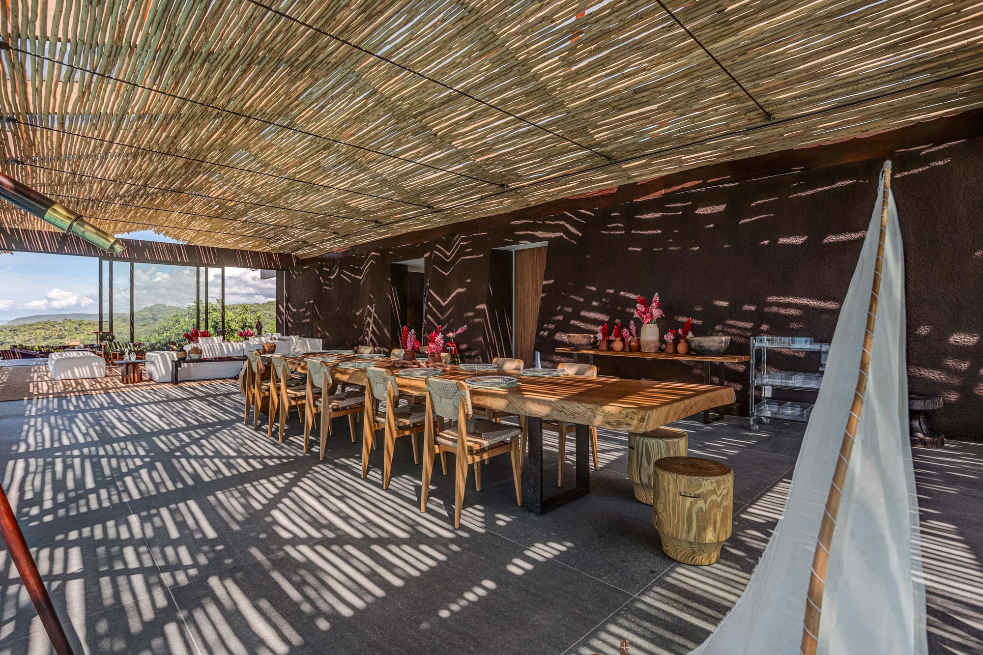Long timber dining table and chairs on a concrete floor striped with sunlight filtering through the cane ceiling, beside a dark rammed wall