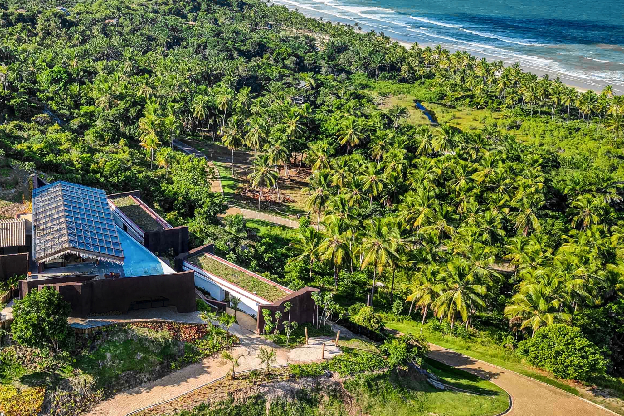 Oblique aerial view of the glass-roofed pavilion, green roofs and winding paths among dense coconut palms, with surf along the shore