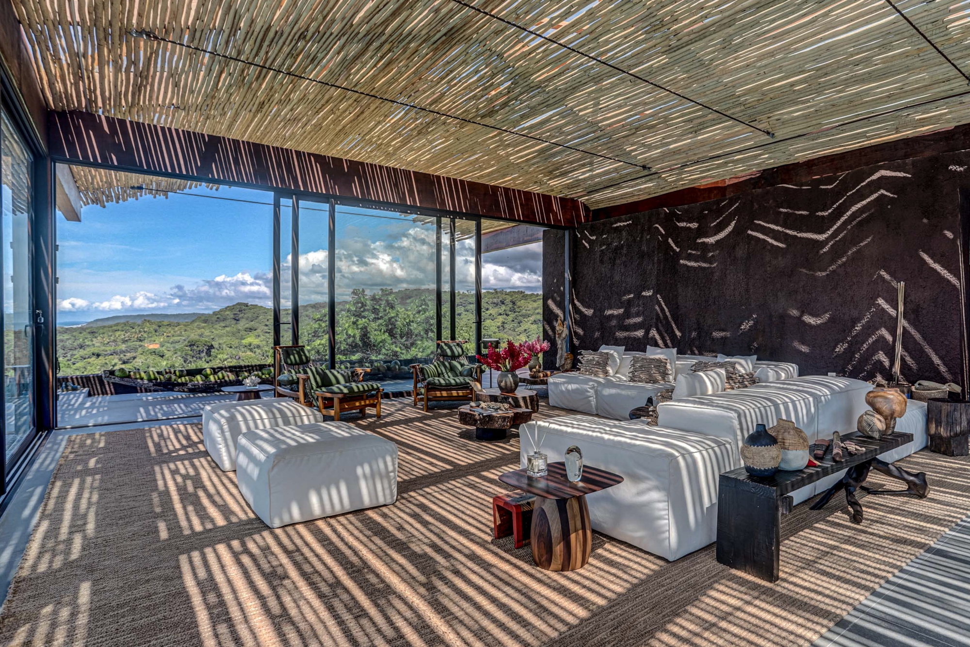 Lounge with white sofas under a woven reed ceiling, striped shadows across the jute rug and forested hills beyond the glass