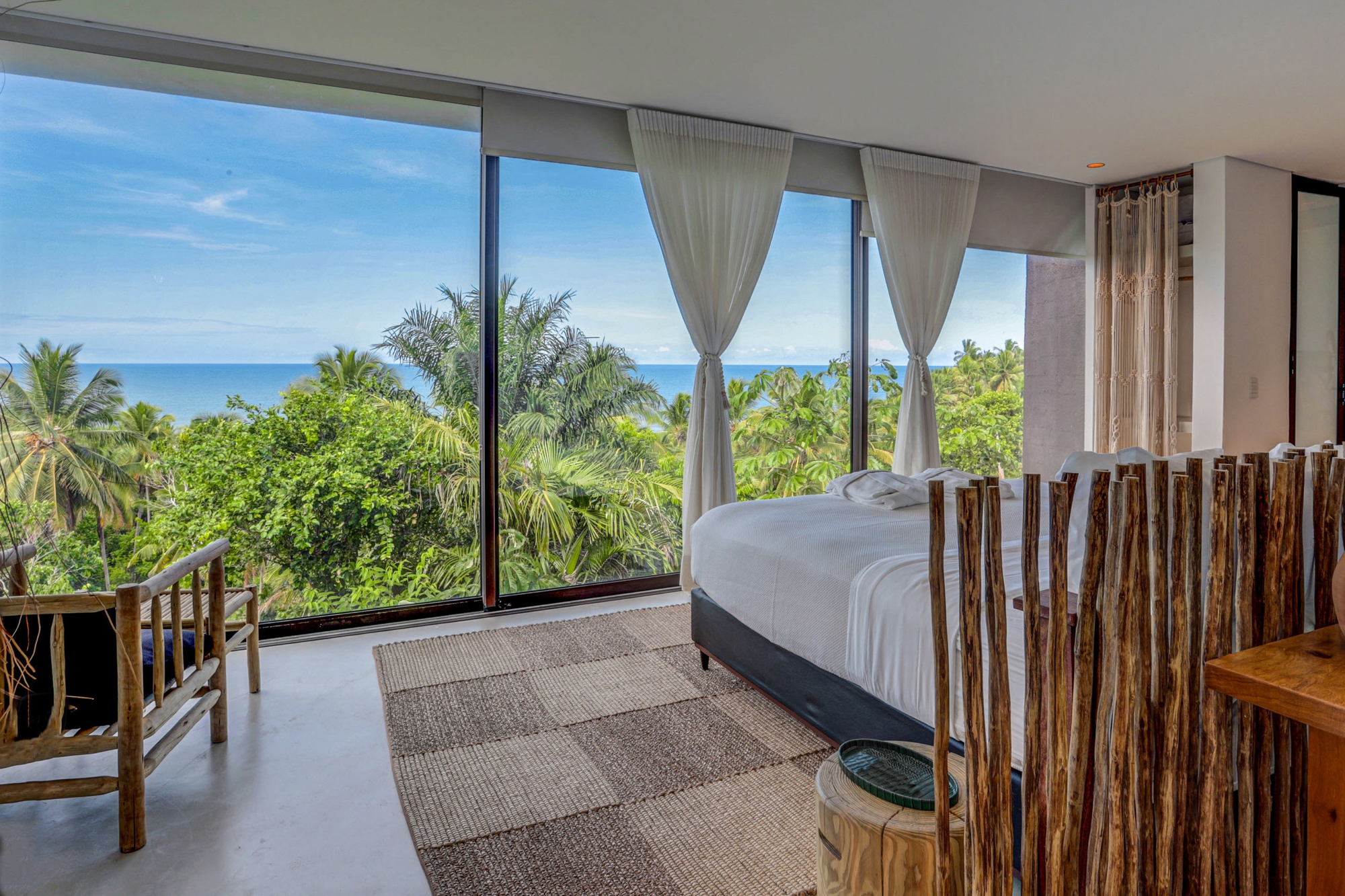 Bedroom with floor-to-ceiling glazing, linen curtains and a branch headboard screen facing palm canopy and ocean