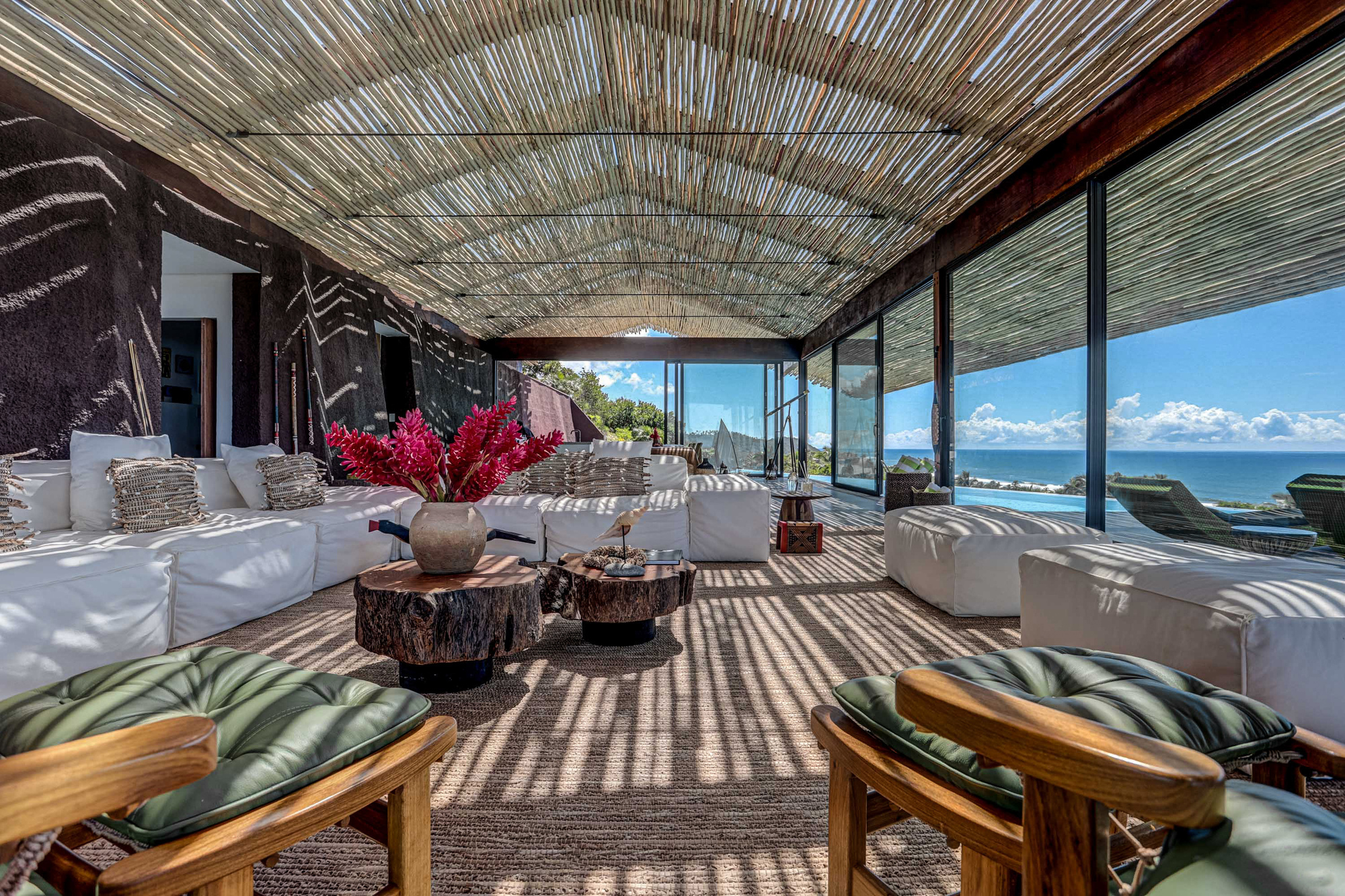 Open pavilion with white sofas, stump tables and green-cushioned teak chairs under a vaulted reed ceiling in afternoon sun