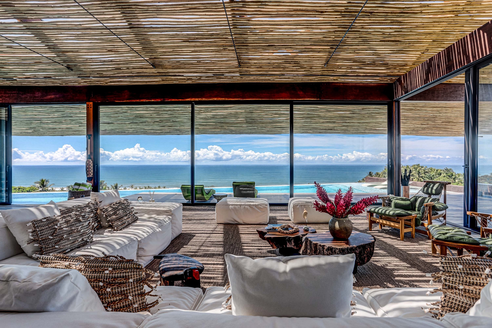 Seating area facing sliding glass walls, with a red ginger flower vase on a log table and ocean horizon beyond