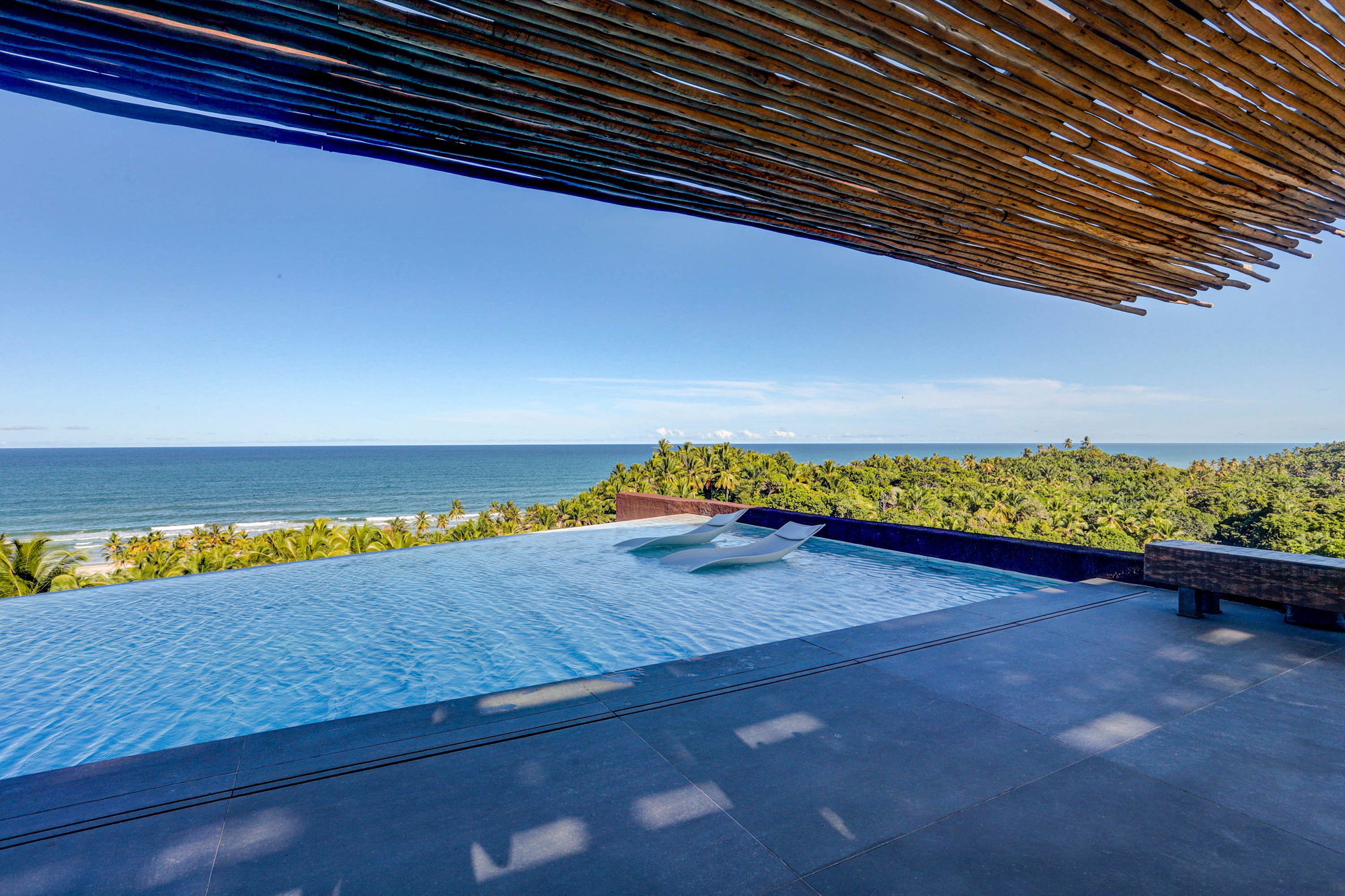 Infinity pool terrace with two white loungers beneath the reed roof edge, overlooking palm forest and sea under clear sky