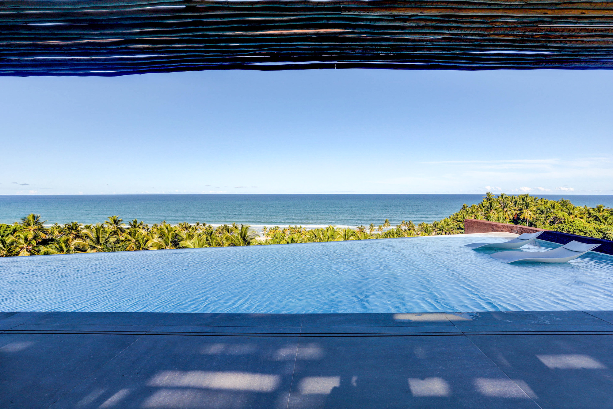 Infinity pool edge under a woven timber ceiling, with two loungers facing palm canopy and the ocean in clear midday light