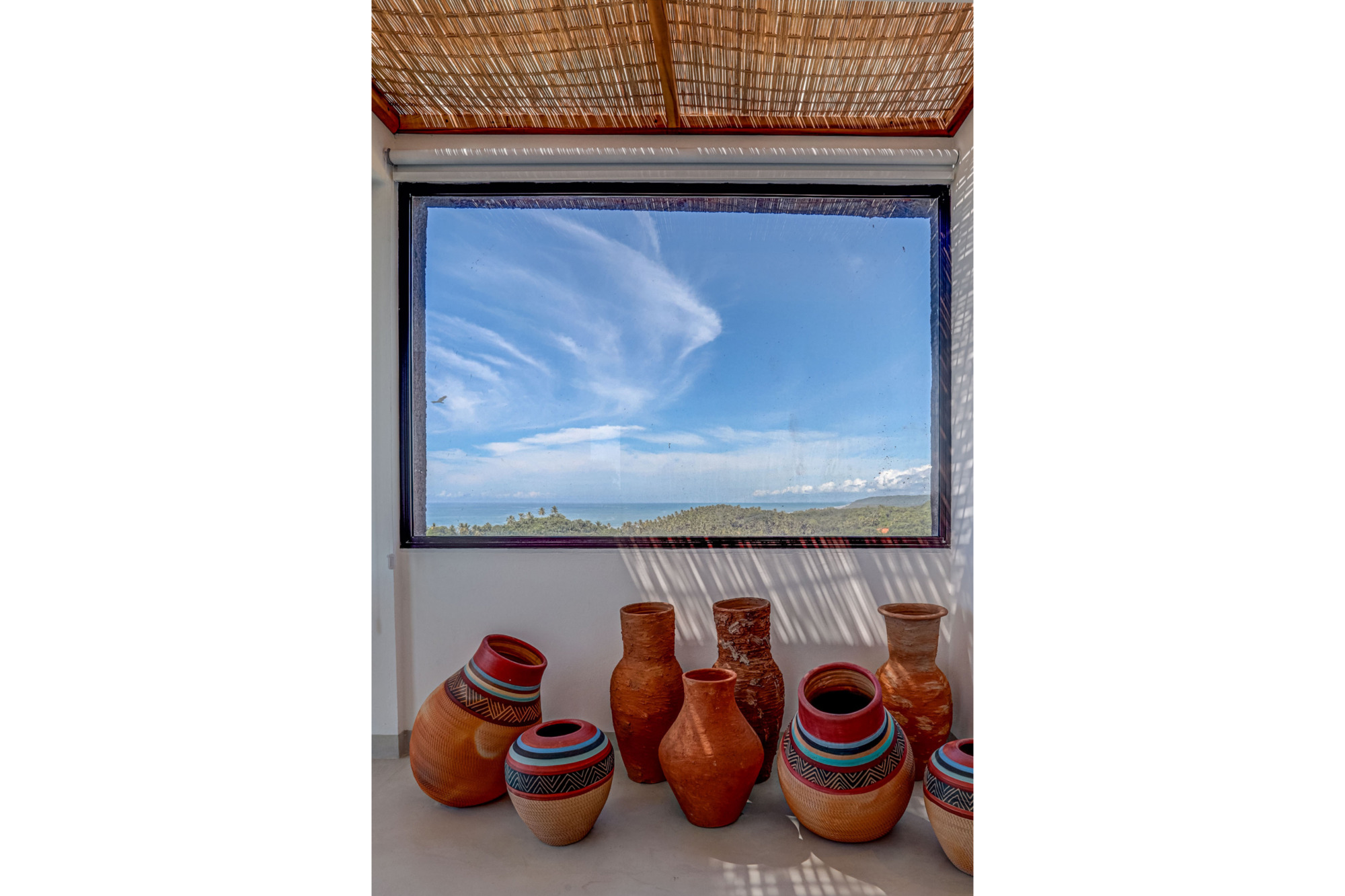 Painted terracotta vessels lined along a wall beneath a framed window of sea and sky, with a reed ceiling above