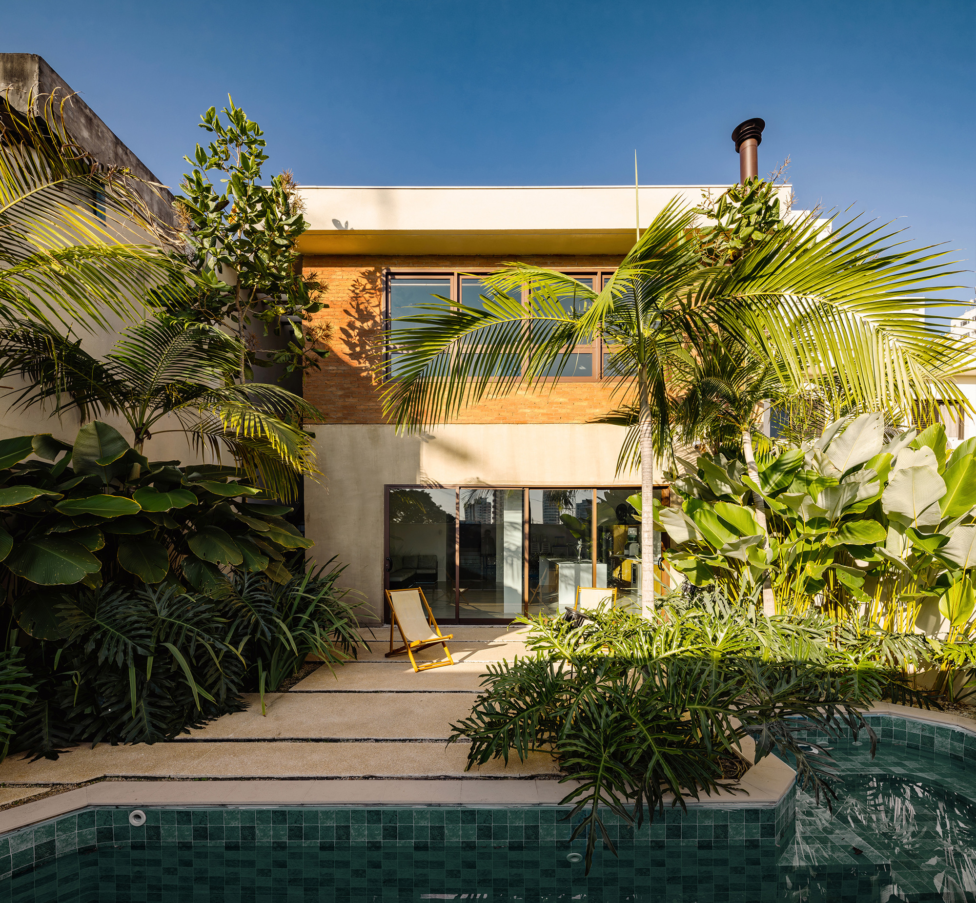 Rear garden facade with brick upper volume and glazed doors behind a green-tiled pool and palms in late sun