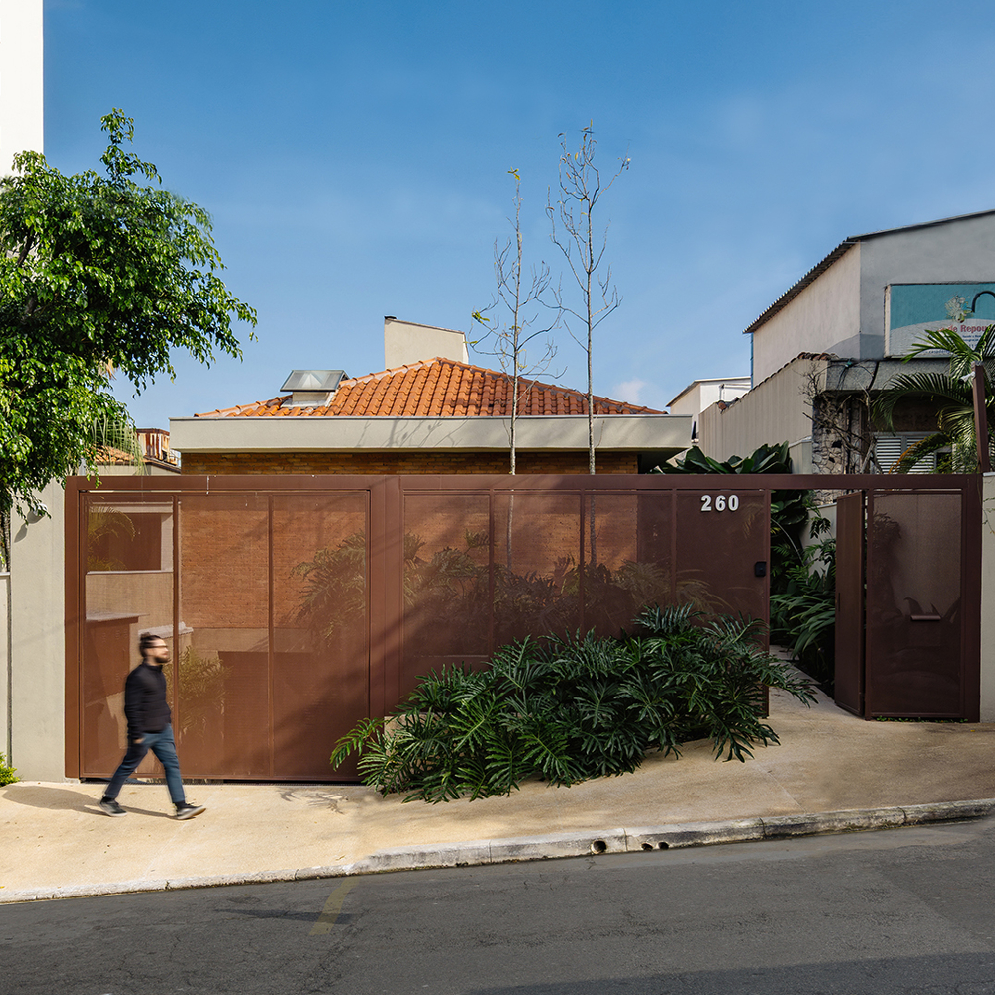 Street frontage with rusted steel perforated gate hiding a clay-tiled roof, as a man walks past under clear sky