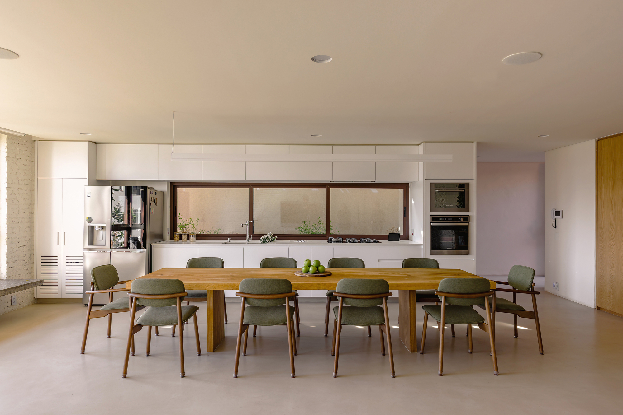 Open kitchen with long timber dining table, green upholstered chairs and a strip window above the white counter in soft daylight