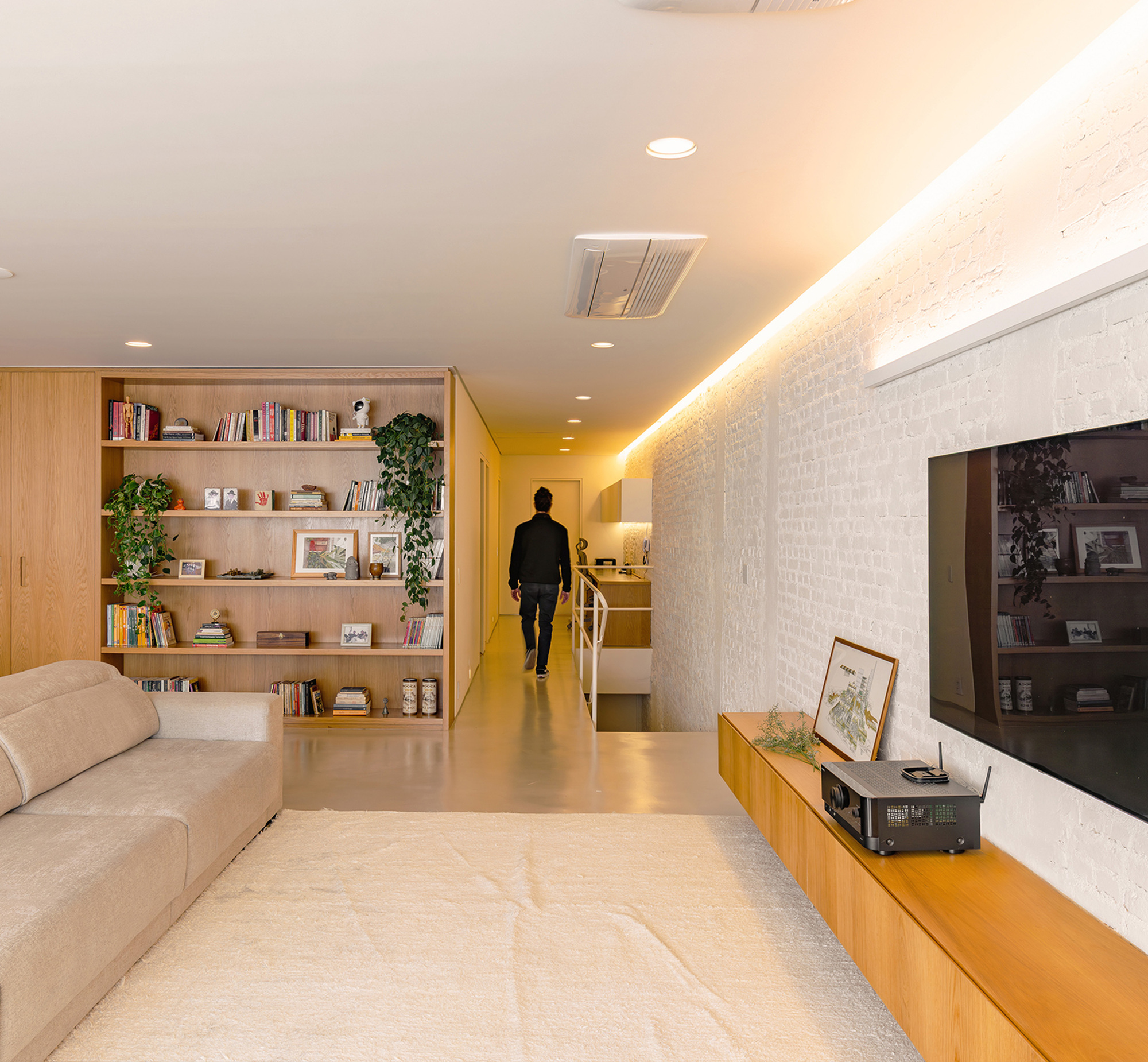 Living room with oak bookshelf, white brick wall lit by a cove strip, and a man walking down the corridor